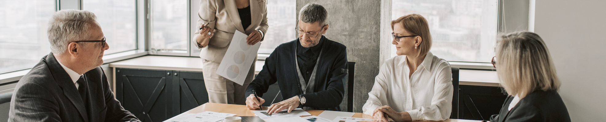 Five people sit at a conference table overlooking a city and have papers with pie charts.