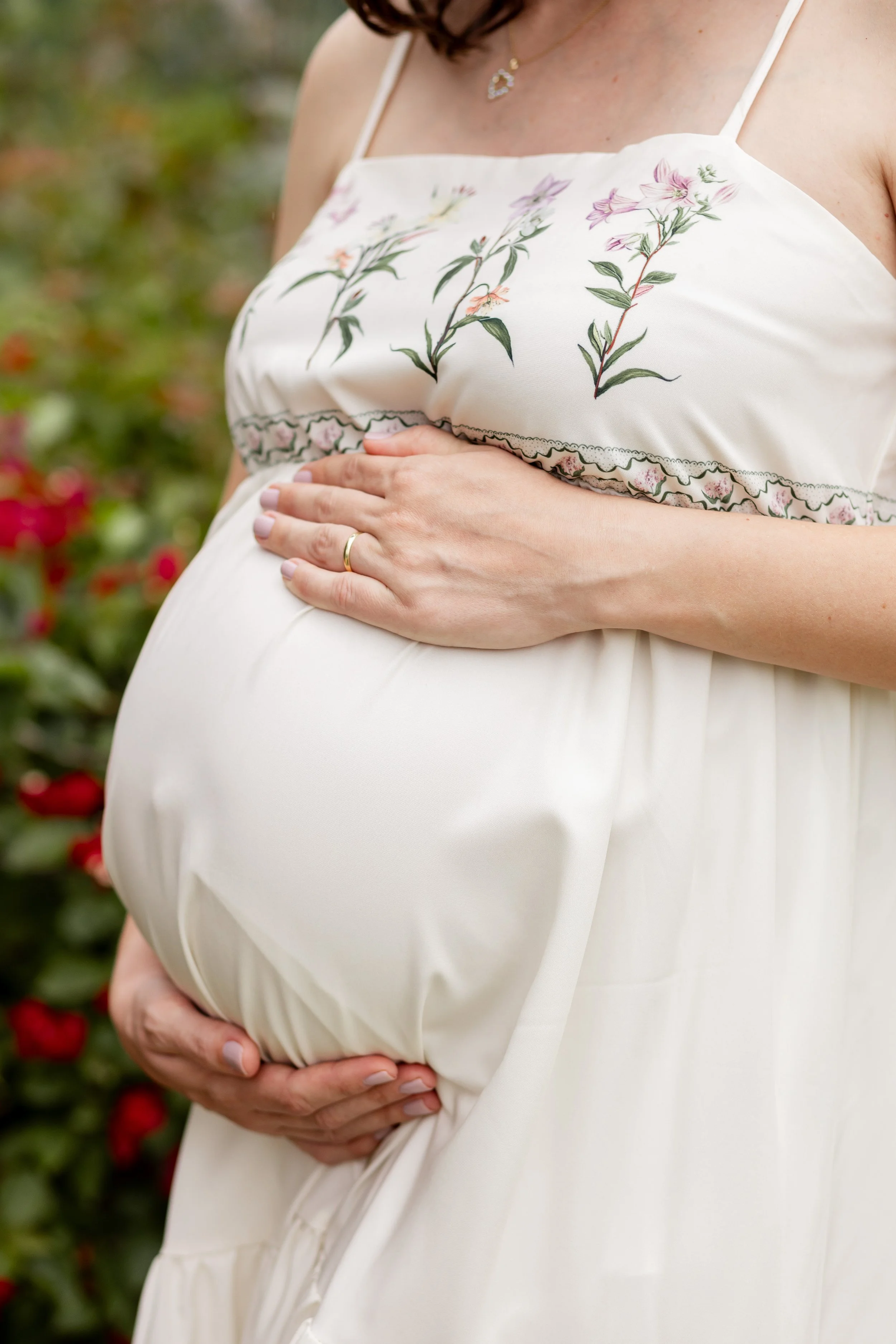 Mulher grávida com vestido branco com detalhes florais, segurando sua barriga com as mãos na frente do corpo.