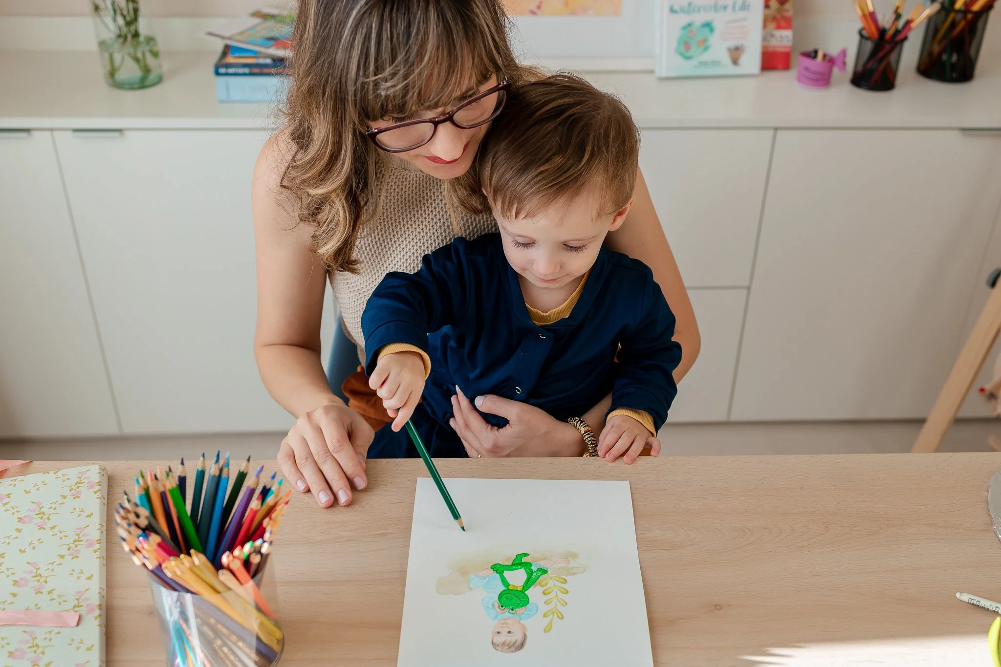 Uma professora ajudando um menino a fazer um desenho com lápis de cor na mesa de uma sala de aula, com diversos materiais escolar ao redor.