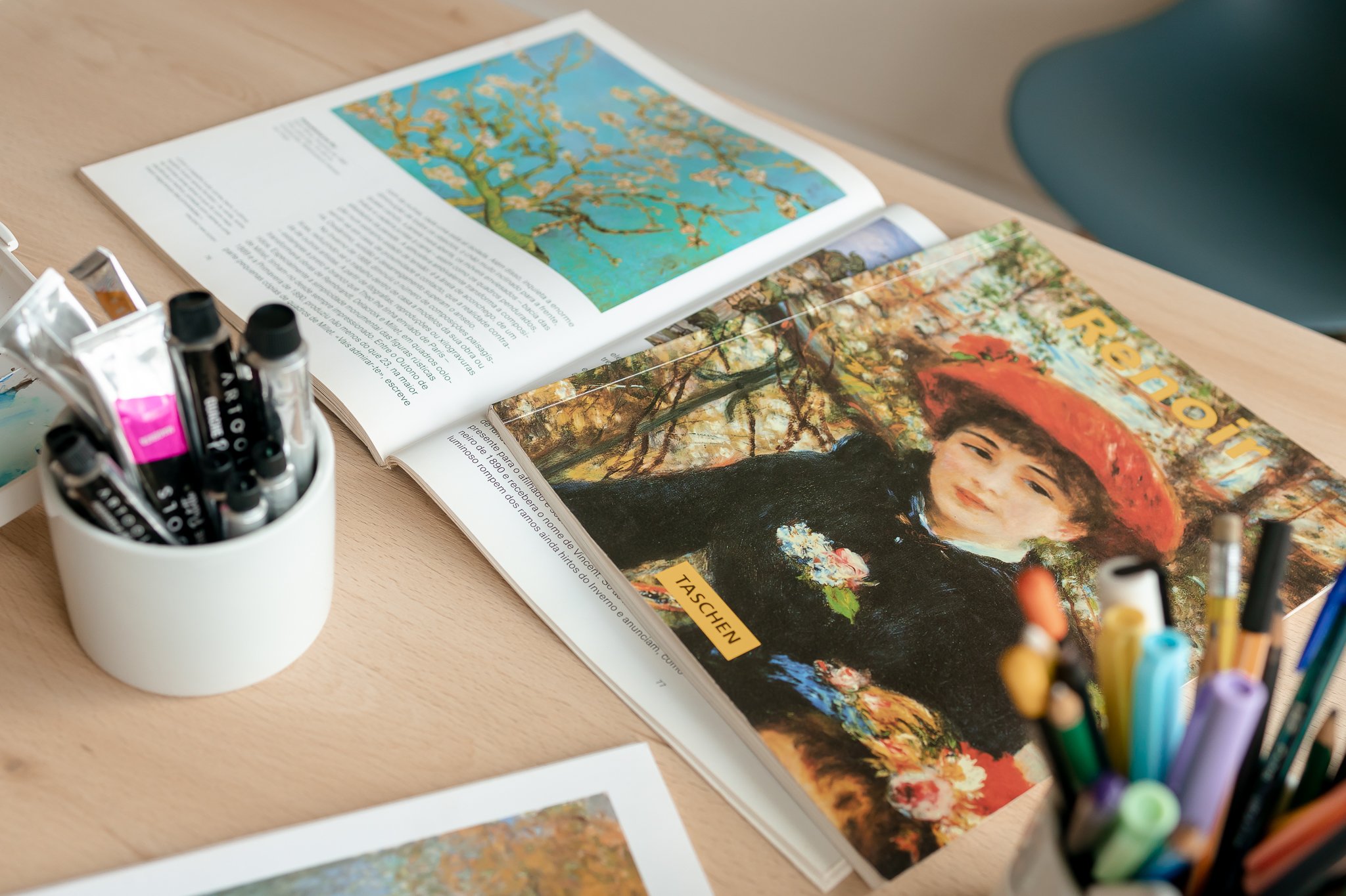Revistas abertas sobre a mesa, uma exibindo uma pintura de árvore com flores e texto e outra com uma foto de uma mulher de chapéu floral ao ar livre. Ao lado, um recipiente com marcadores coloridos.