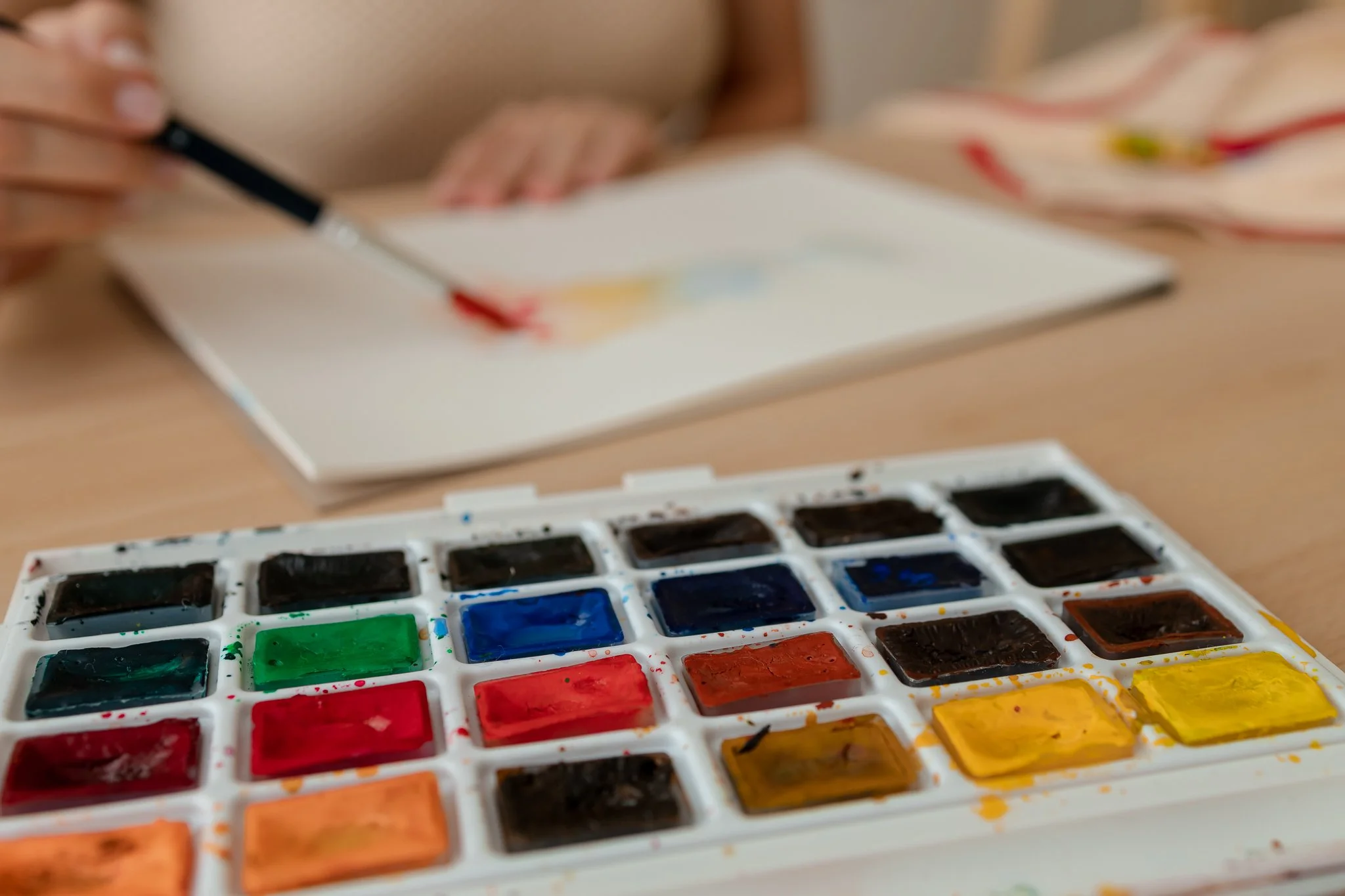 Paleta de tintas com várias cores, incluindo verde, vermelho, azul e amarelo, ao lado de um caderno e uma pessoa usando uma caneta ou pincel.