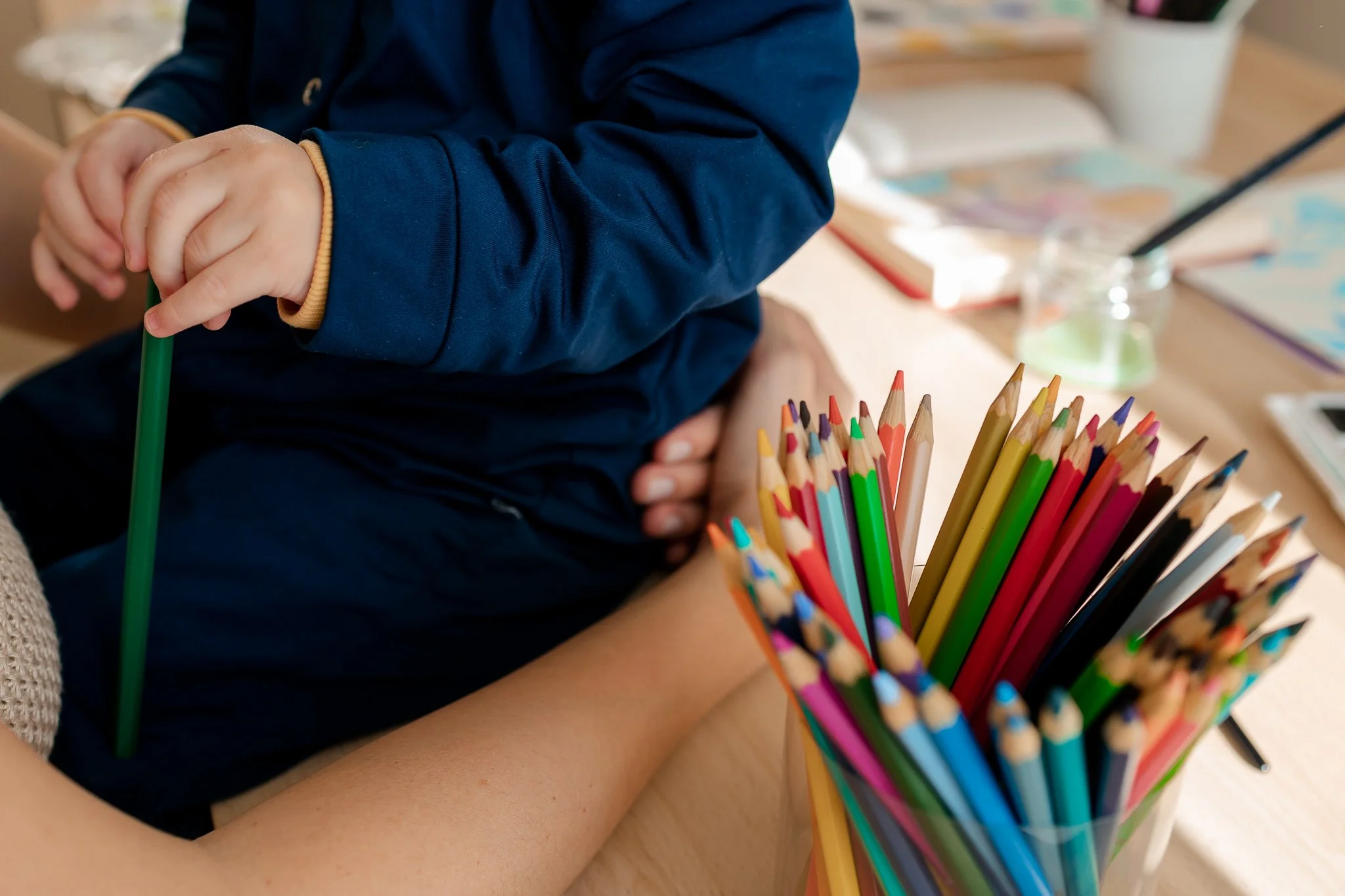 Mãos de criança segurando um lápis verde, próximo a uma caixa com lápis de cor coloridos em um ambiente de sala de aula.