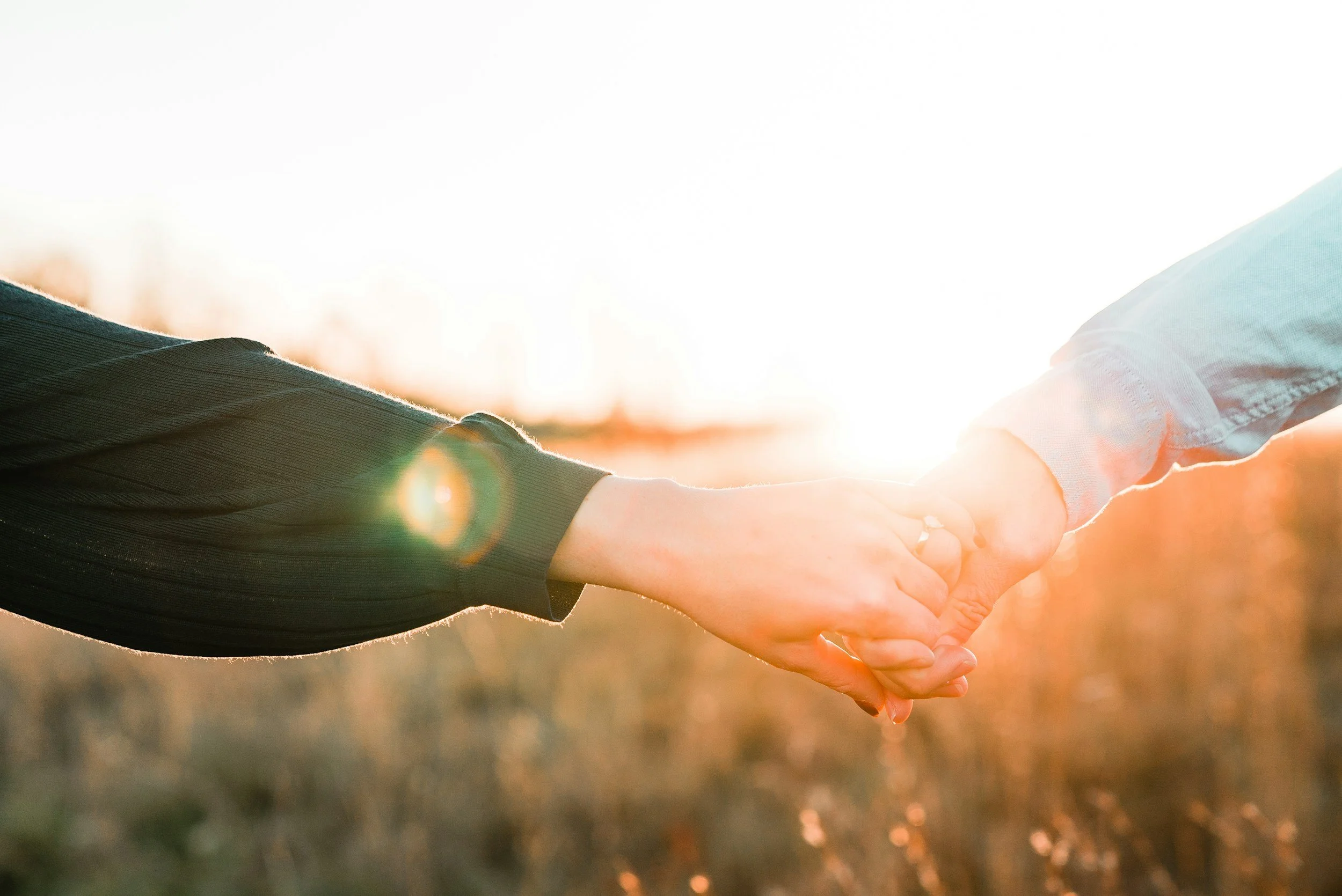Two people holding hands at sunset or sunrise with warm sunlight and a blurred natural background.