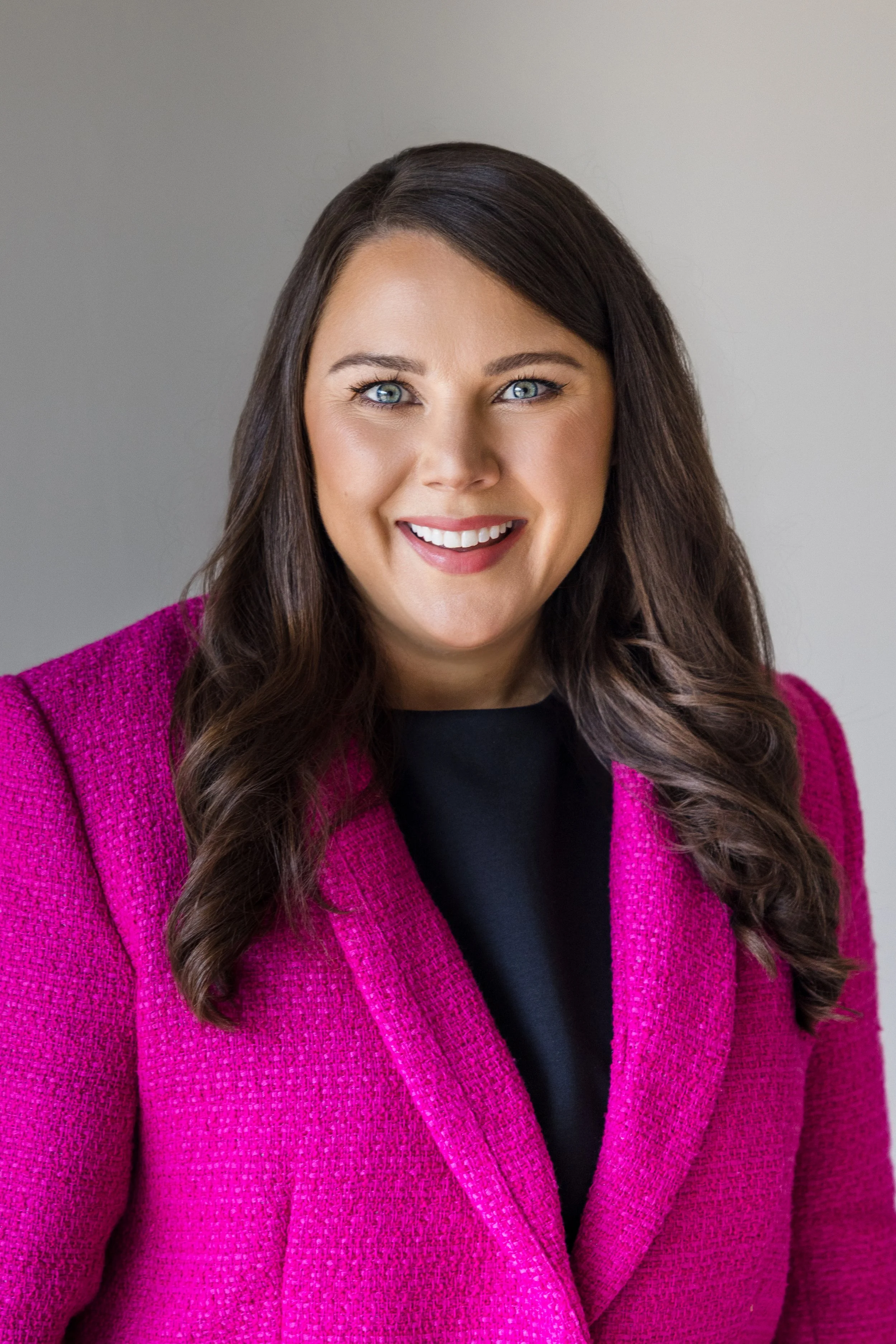 A woman with long wavy brown hair, light skin, and blue eyes smiles in a portrait. She wears a bright pink textured blazer over a black top, against a plain, light gray background.