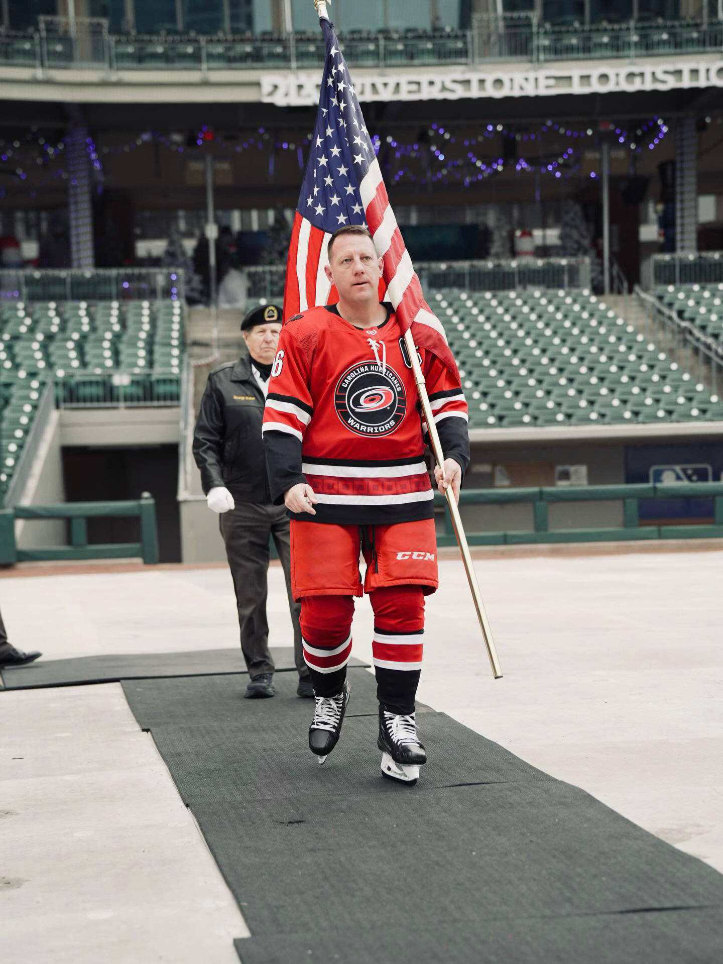 A man in a red hockey uniform and skates holding an American flag walks on a black mat on a skating rink, with a woman behind him also in a hockey uniform, and empty seating in the background.