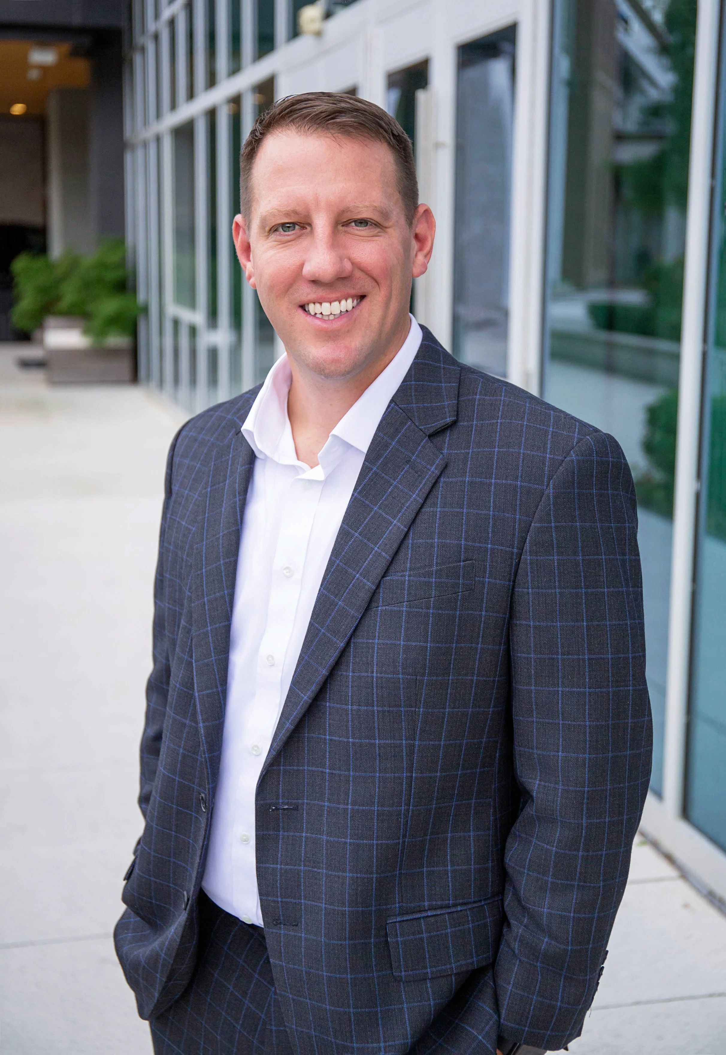 A man with short brown hair and blue eyes dressed in a dark blue plaid suit and white shirt, standing outside near a modern building with large glass windows and greenery in the background.