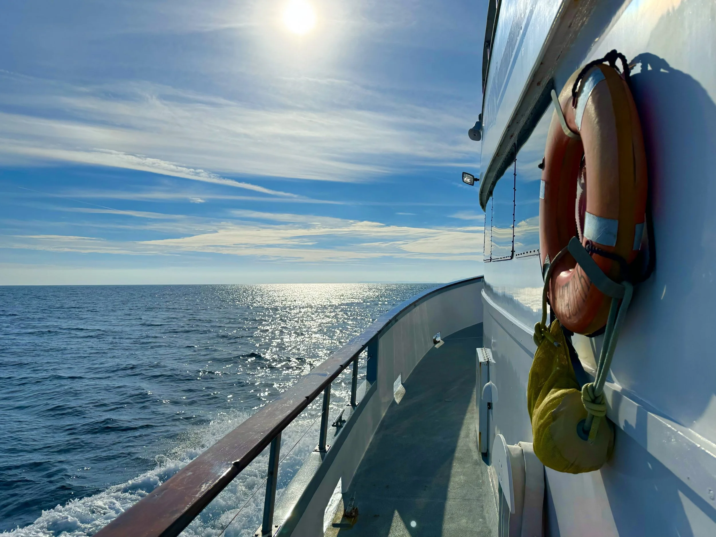 View from the side of a boat showing the ocean, partly cloudy sky, and the sun shining above the horizon. A life jacket is hanging on the boat's railing.