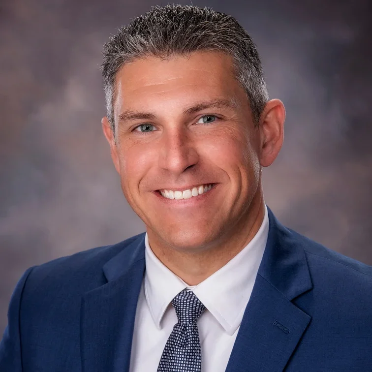 Professional headshot of a smiling man in a navy blue suit, white shirt, and patterned tie, with short gray hair and a neutral background.