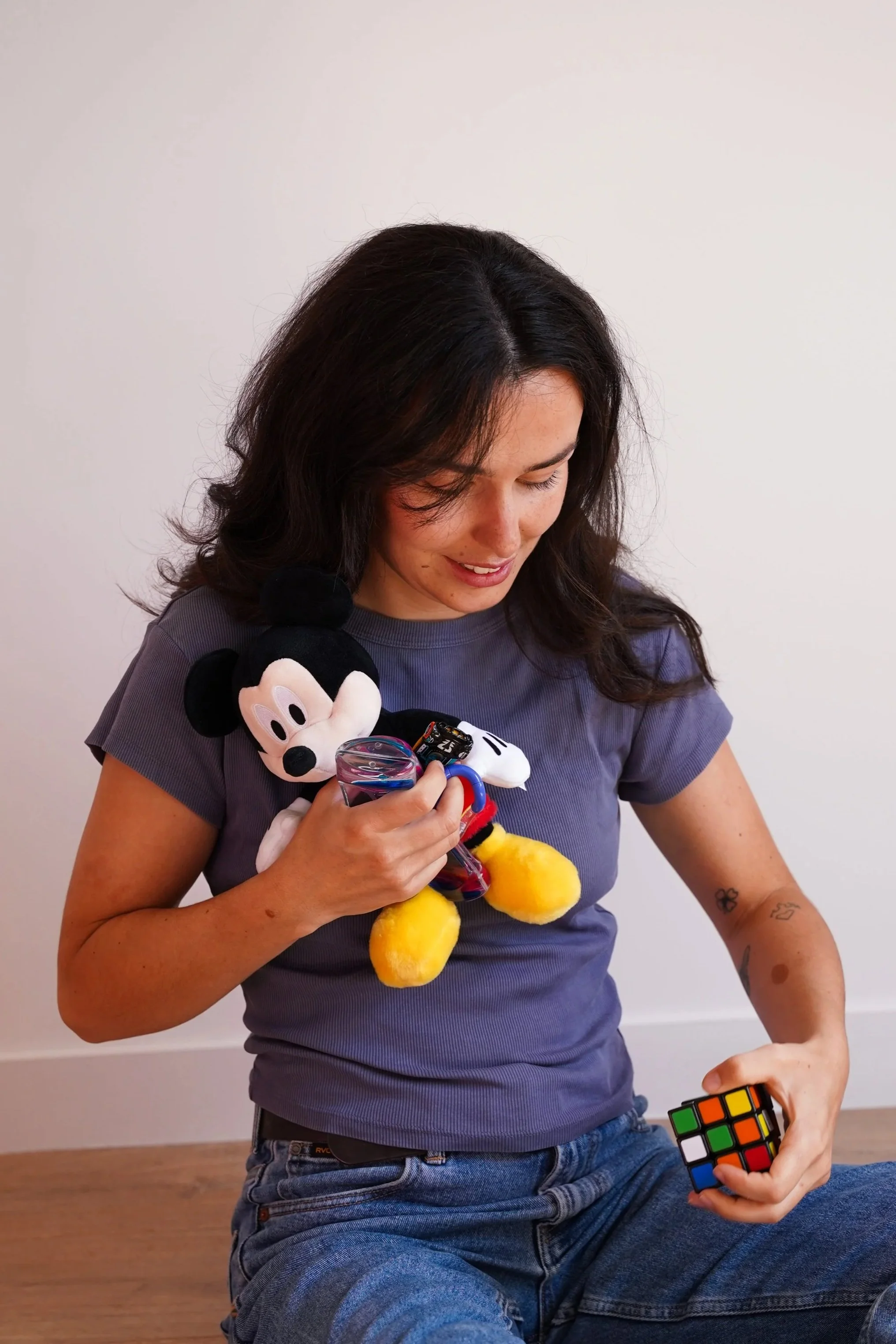 Une femme assise sur le sol, tenant une figurine de Mickey Mouse dans la main gauche, regarde un Rubik's Cube dans la main droite, portant un t-shirt gris et des jeans, avec un mur blanc en arrière-plan.