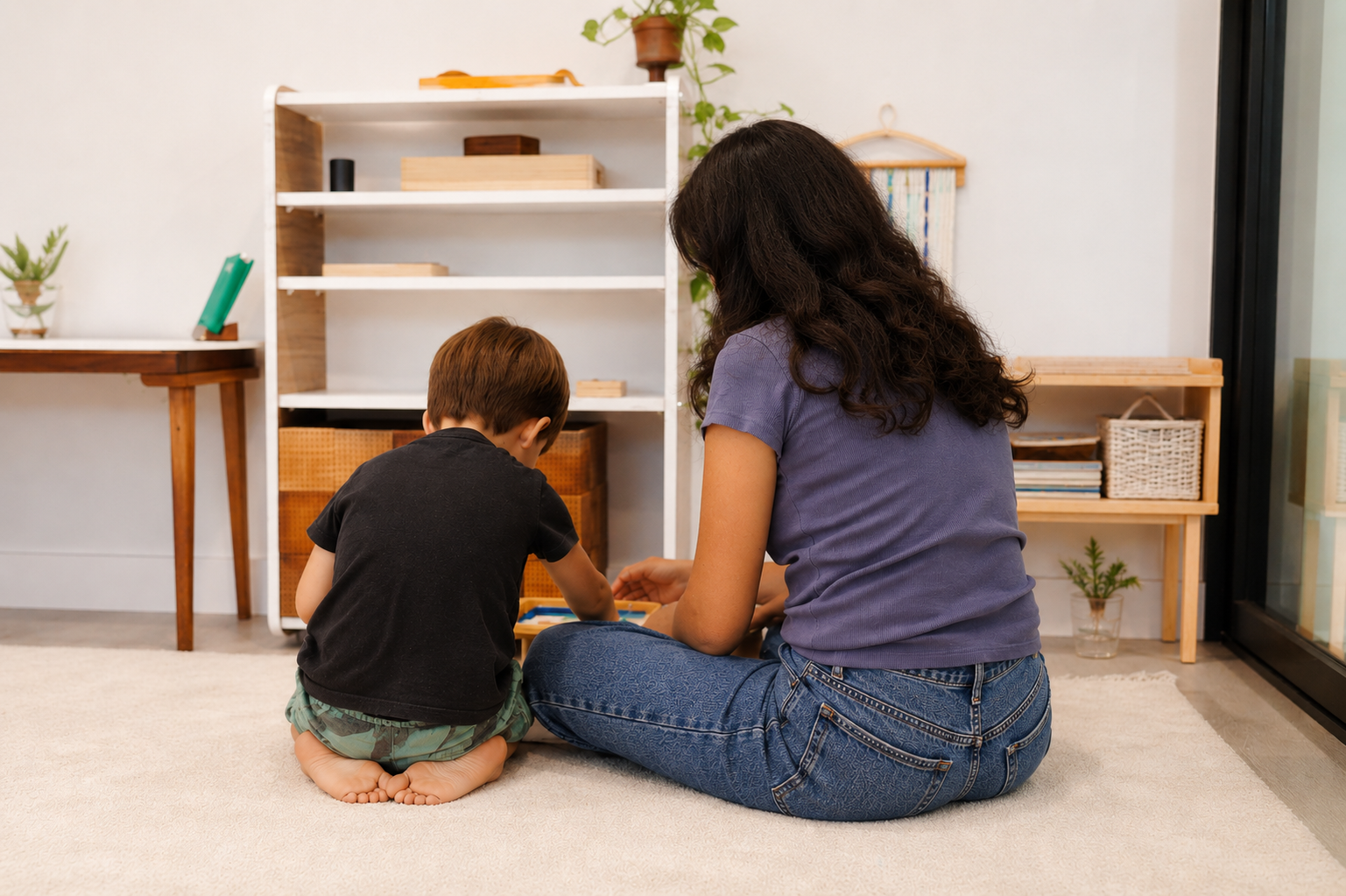 Une femme et un jeune garçon assis par terre jouent à un jeu de société dans un salon. Il y a une étagère blanche et des plantes en arrière-plan, avec des meubles en bois autour.