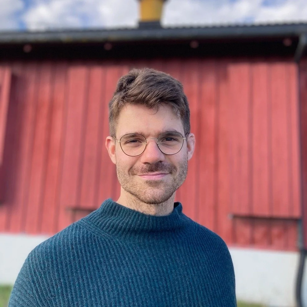 A young man with brown hair, glasses, and a beard wearing a blue sweater standing in front of a red wooden building.
