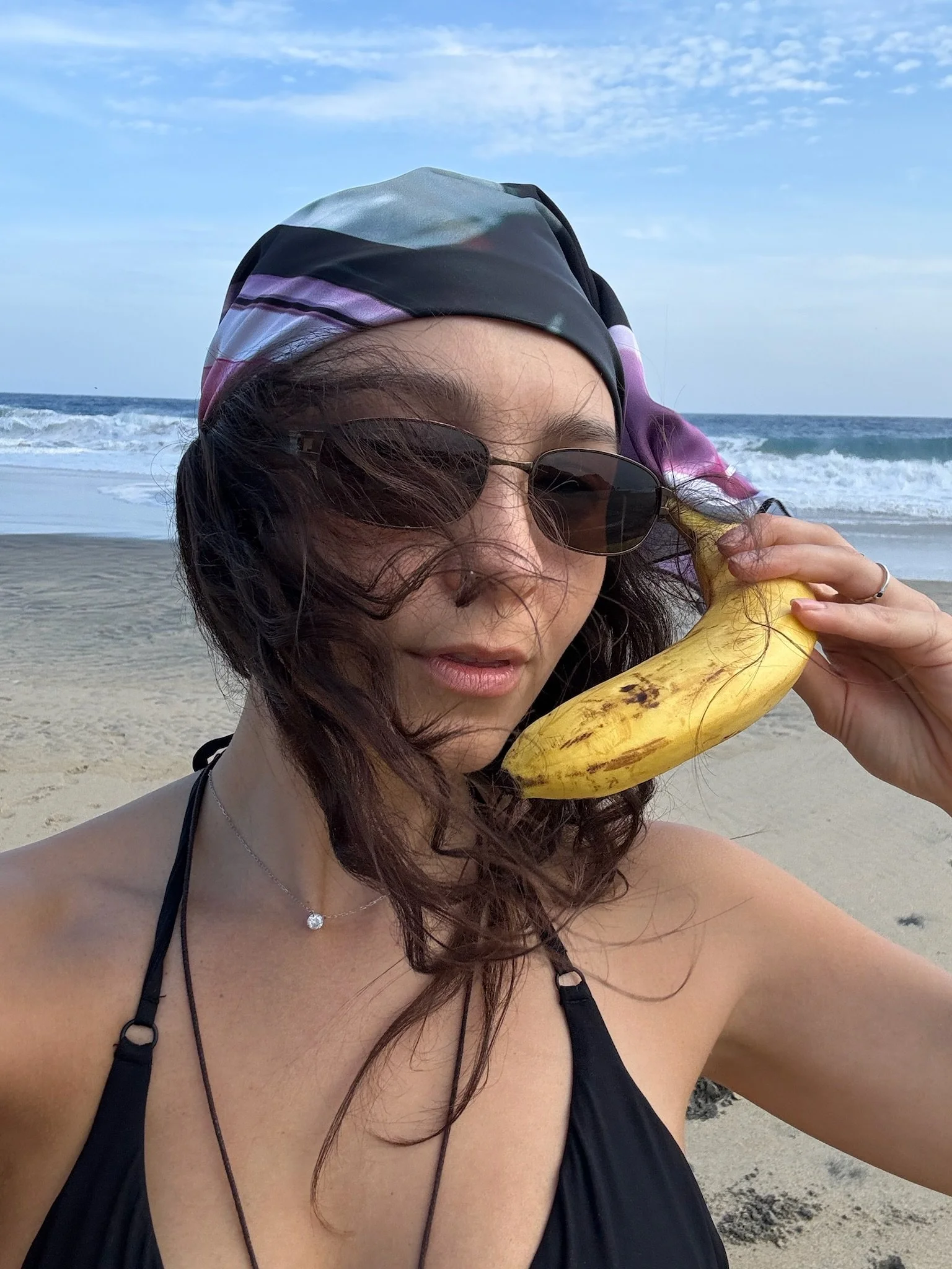 A woman with wavy brown hair wearing sunglasses, a black top, and a scarf on her head, holding a banana, at the beach with sand, ocean waves, and blue sky in the background.