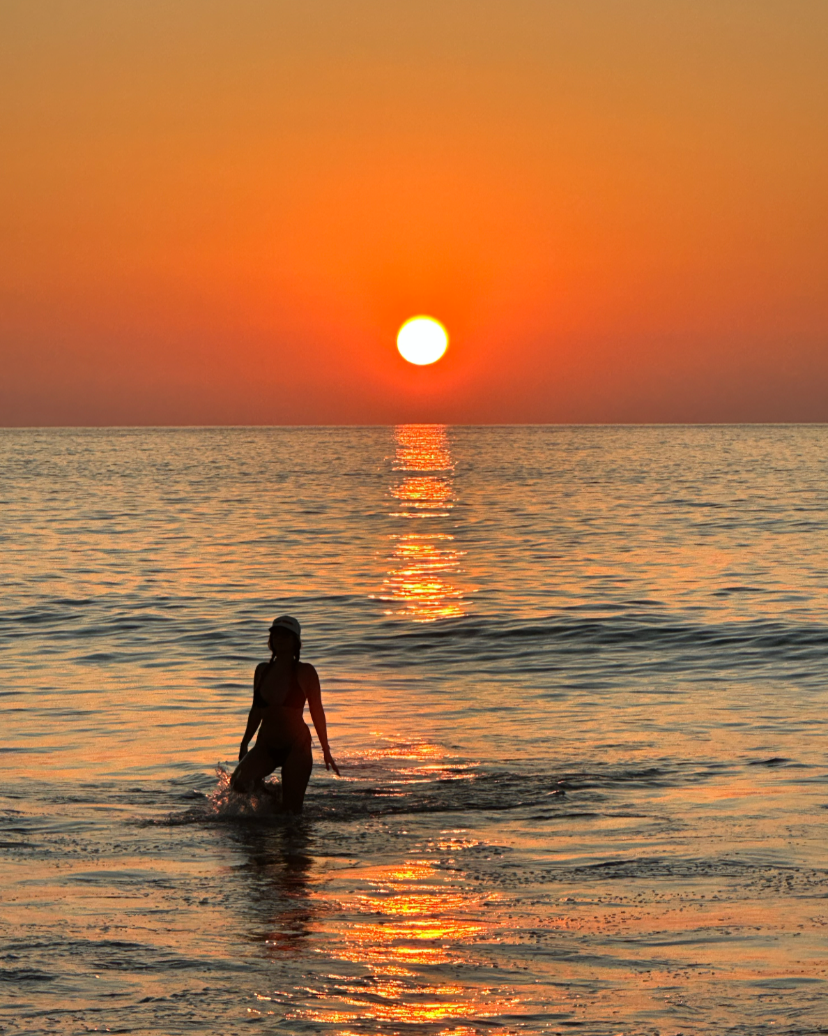 Silhouette of a woman standing in the water at sunset, with the sun low in the sky and colorful sky reflected on the ocean.
