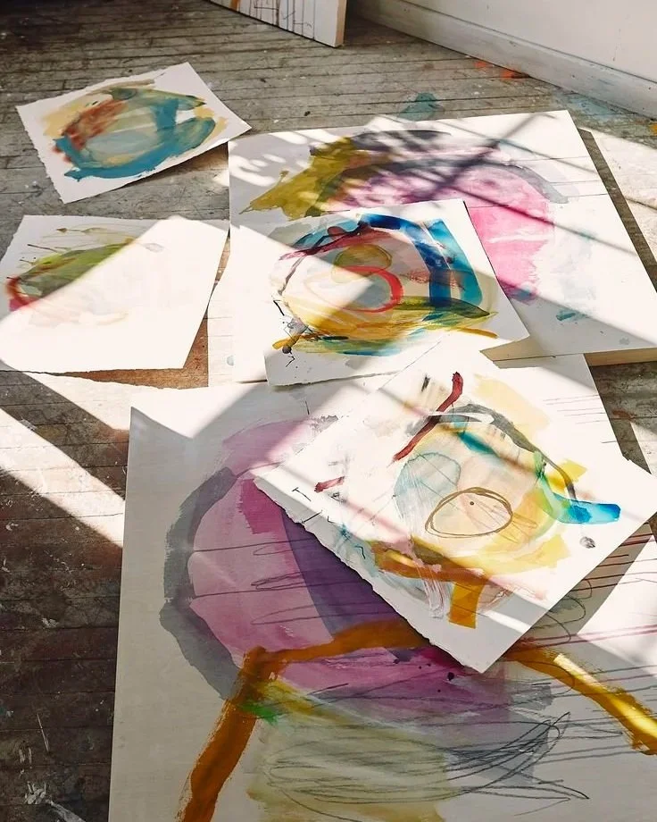 Several sheets of paper with colorful abstract watercolor paintings scattered on a wooden floor, with shadows cast on them.