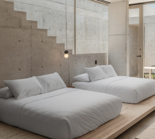 Two neatly made beds with white bedding in a minimalist bedroom with concrete walls and a large window.