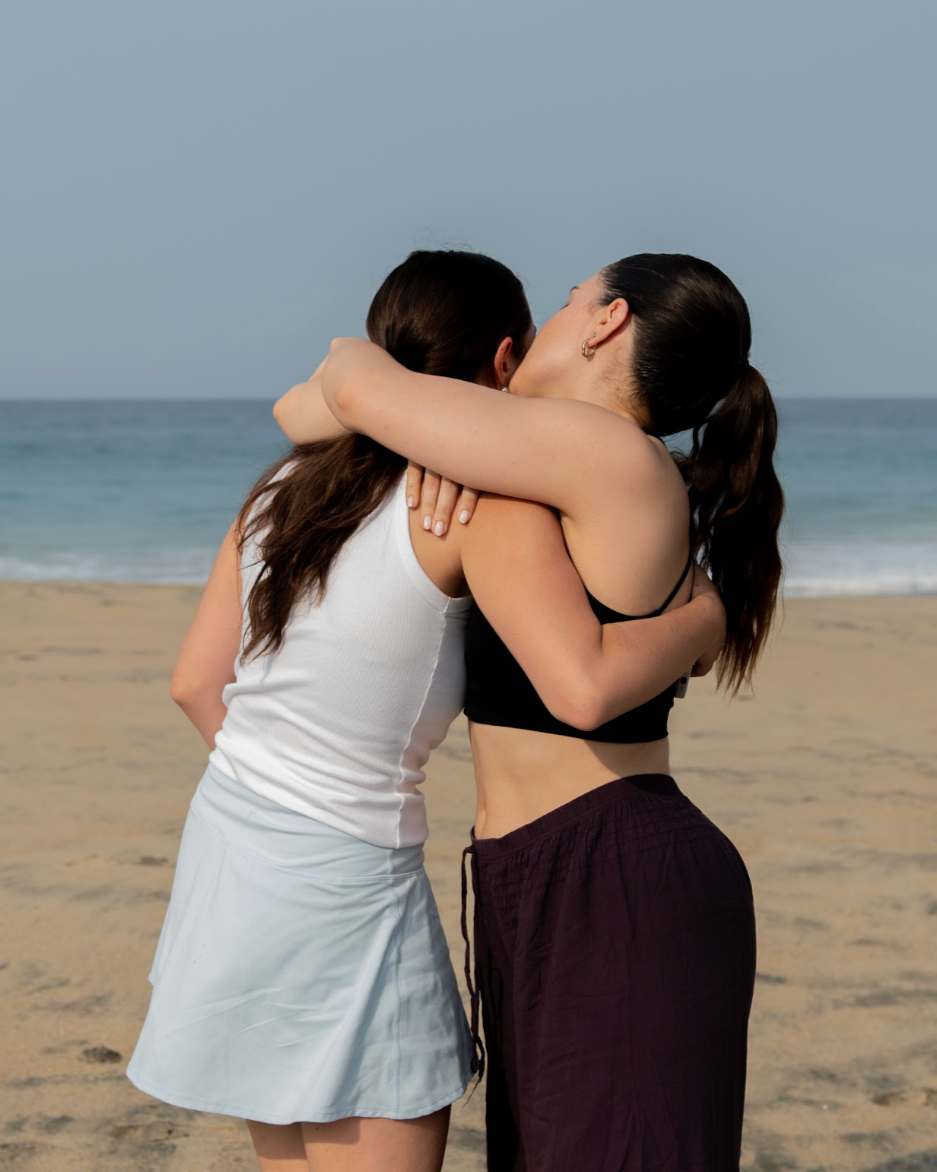 Two women hugging and kissing on the beach with ocean waves in the background.