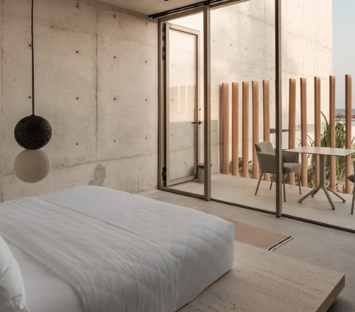 Modern bedroom with concrete walls, a bed with white bedding, a black hanging lamp, and a glass door leading to a balcony with wooden slats and outdoor furniture.