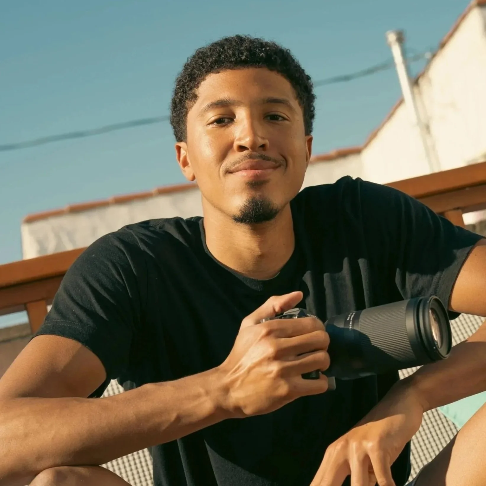 Young man with short curly hair and a goatee smiling and holding a camera, sitting outdoors on a daytime sunny day.