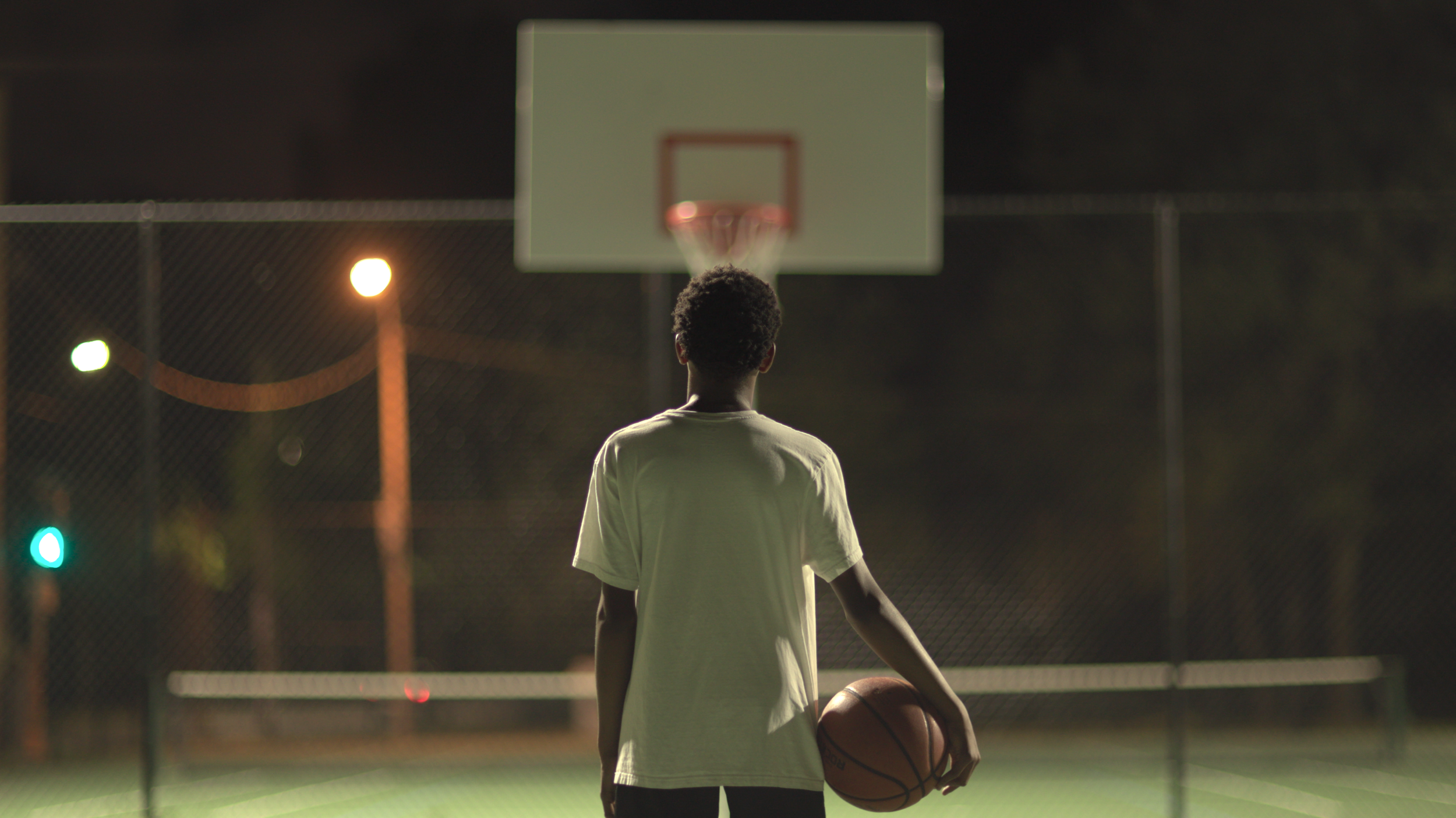 A person standing on a basketball court at night, holding a basketball under their right arm, facing a basketball hoop, with city streetlights visible in the background.
