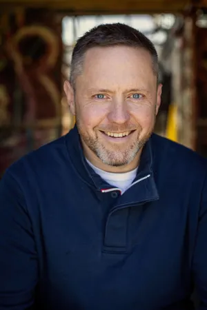 Headshot of a smiling man with short dark hair and a beard, wearing a navy jacket, in an indoor setting with blurred background.