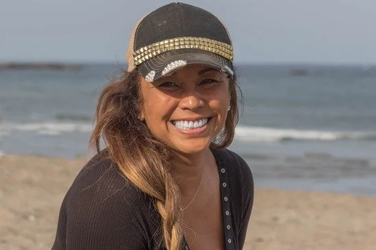 Woman smiling on the beach wearing a black cap and a black top with buttons, ocean in background.