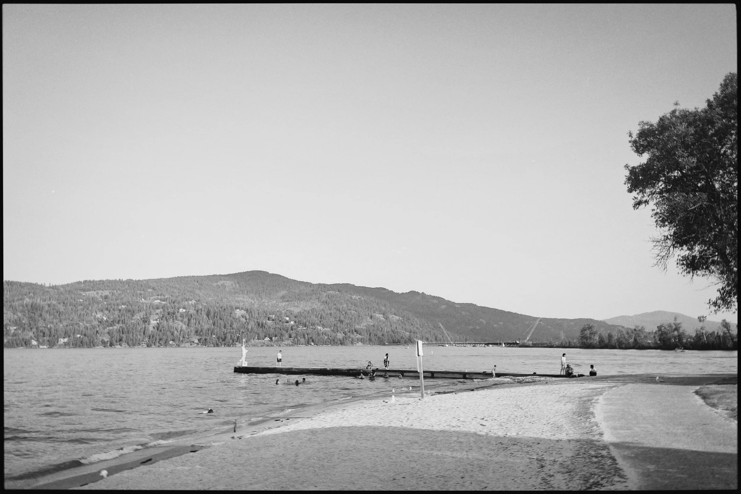 Locals enjoying a summer day at the eastern end of City Beach, swimming and jumping from the iconic Statue of Liberty Pier with Lake Pend Oreille and the surrounding mountains beyond.