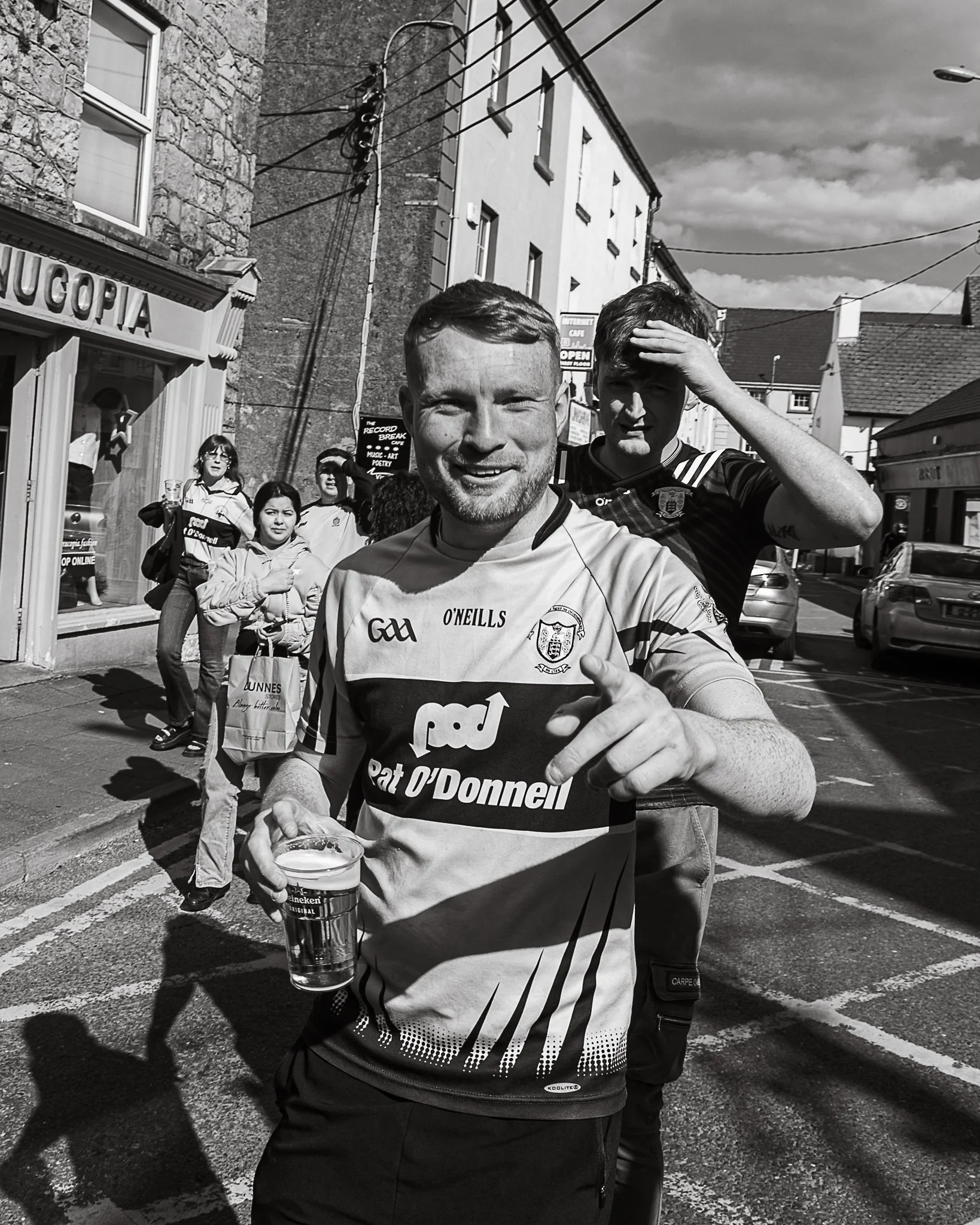 A man in an outdoor urban setting, with one holding a pint glass of beer, and a group of women in the background near a store and a street.