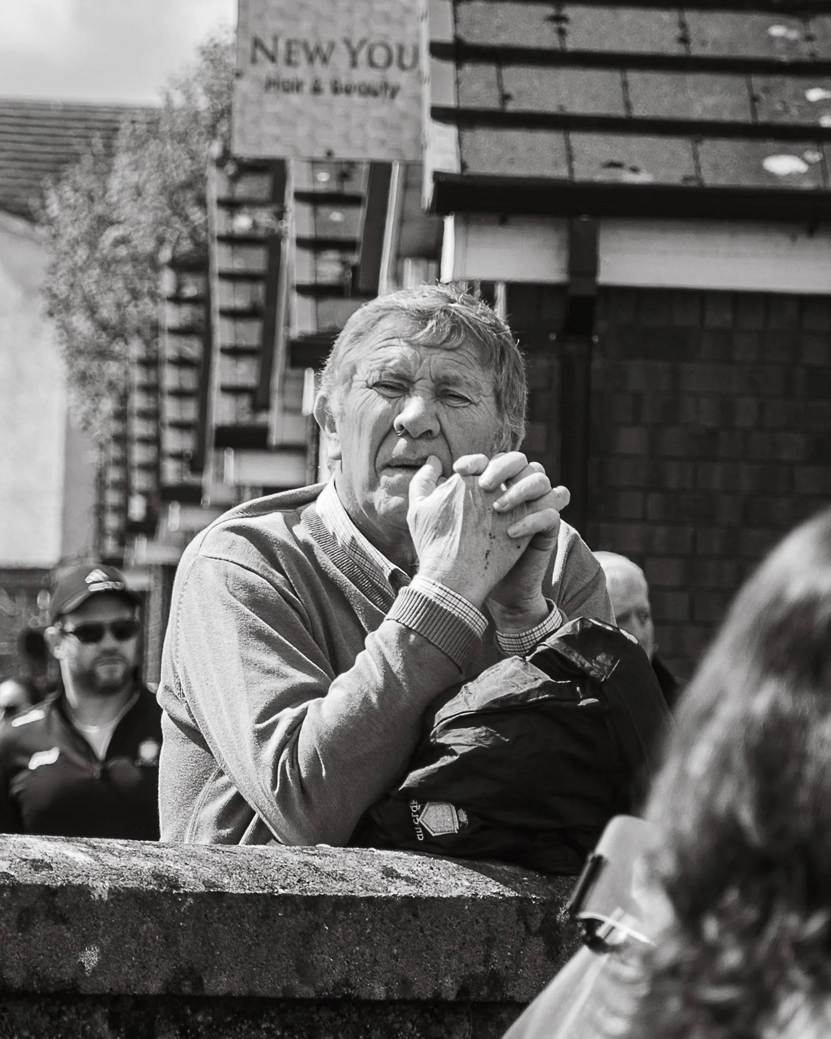 A black-and-white photo of an elderly man with short, light-colored hair, sticking his tongue out and making a face, sitting outdoors at a table. In the background, there is a sign that reads 'NEW YOU Hair & Beauty' and some residential buildings.