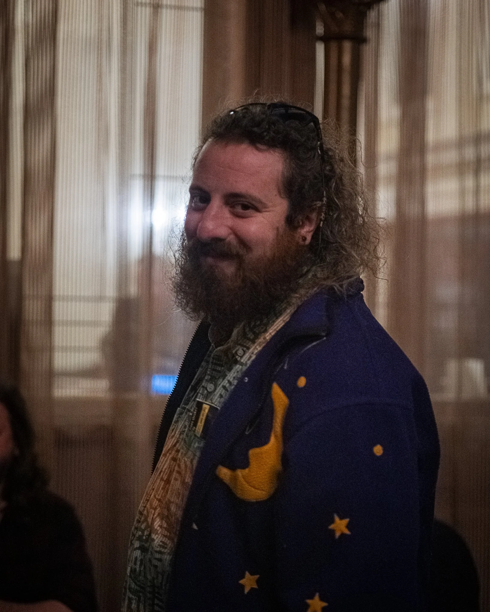 A man with curly hair and a beard, wearing glasses on his head, is smiling slightly. He is dressed in a blue jacket with yellow stars and a plaid shirt underneath. The background consists of vertical blinds with sunlight filtering through.