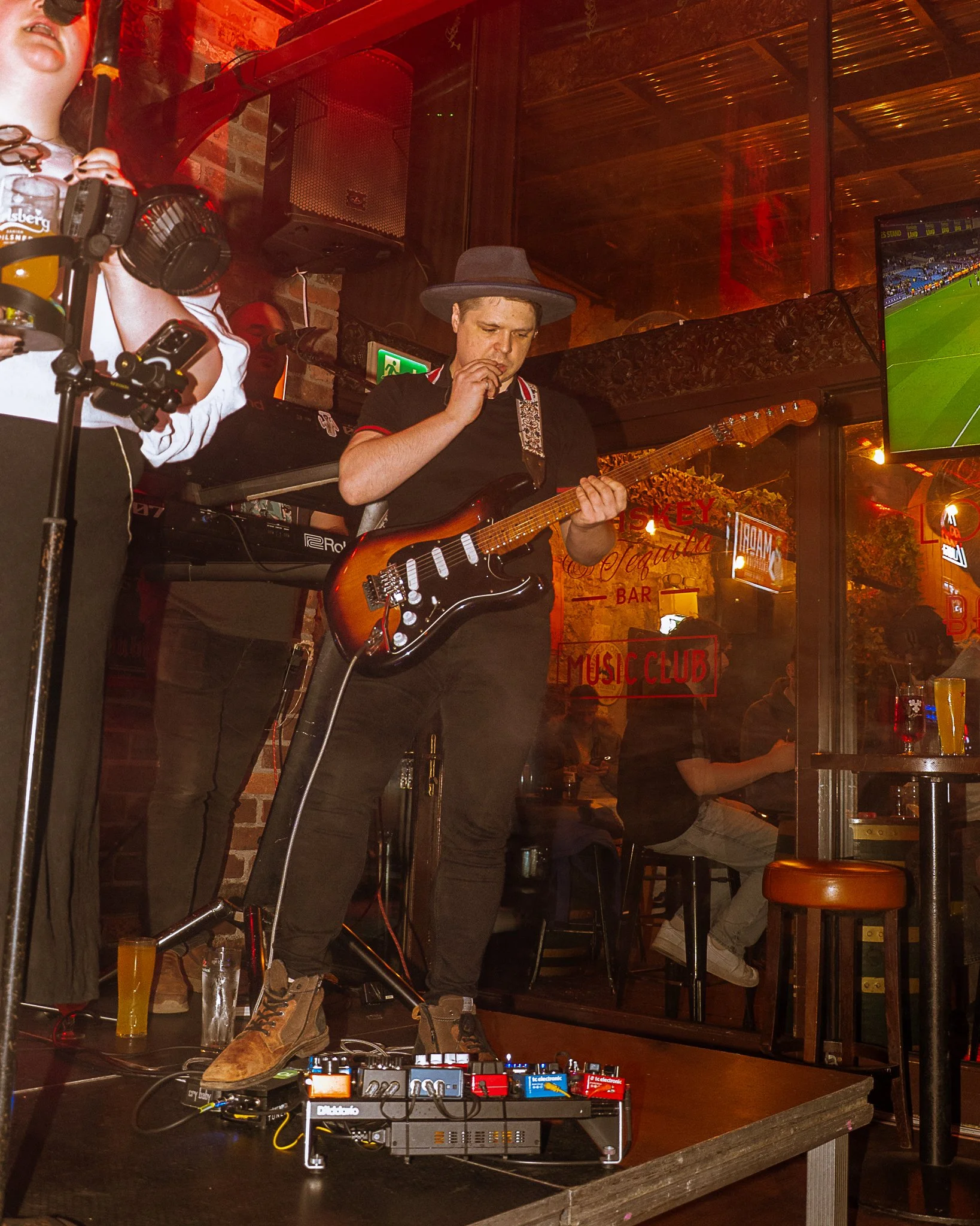 Male musician wearing a hat playing an electric guitar at a music venue. DJ equipment and a beer glass are on the table in front of him. Other people seated inside, visible through the window, and a mounted TV showing a soccer game.