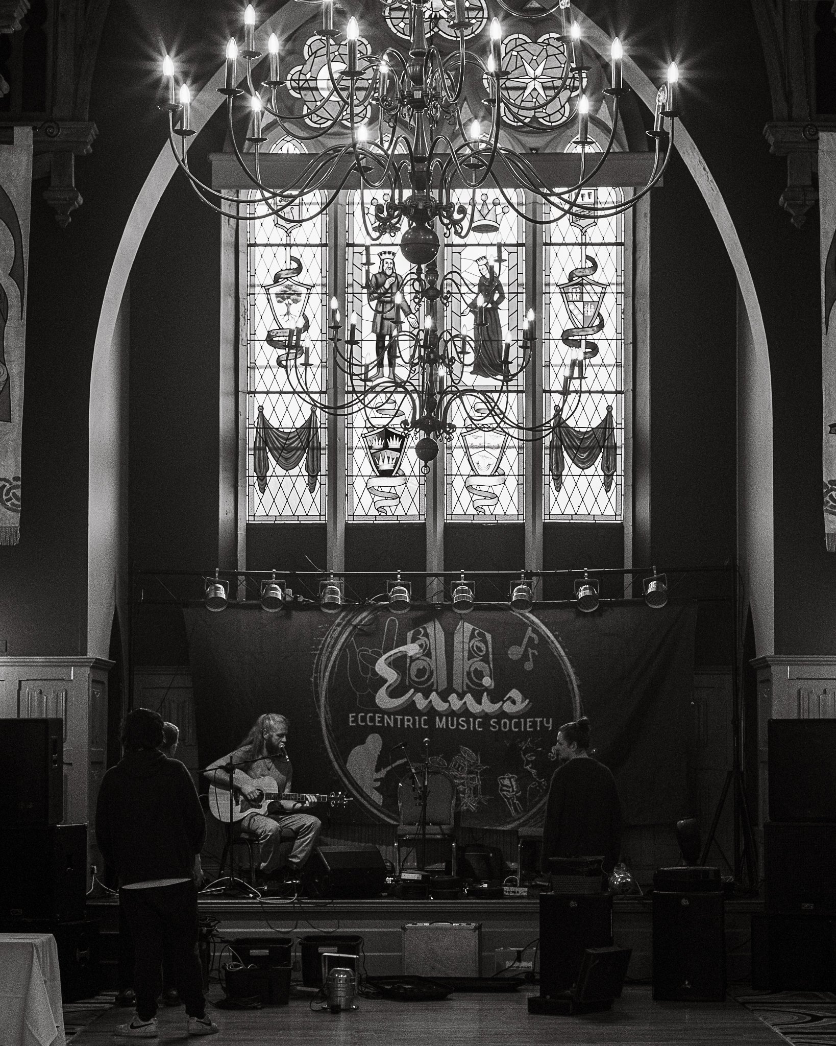 A musician playing an acoustic guitar on stage at the Eccentric Music Society, with a large stained glass window and a chandelier above, and people watching in the audience.