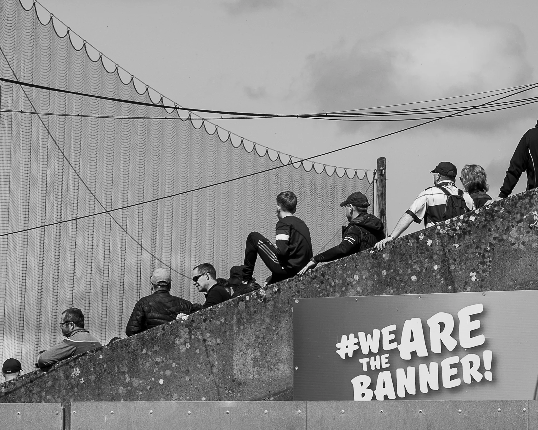 Group of people sitting and standing on a stone wall, facing away, with a large protest sign that reads '#WEARE THE BANNER!' in the foreground. Overhead power lines among clouds are visible in the sky in the background.