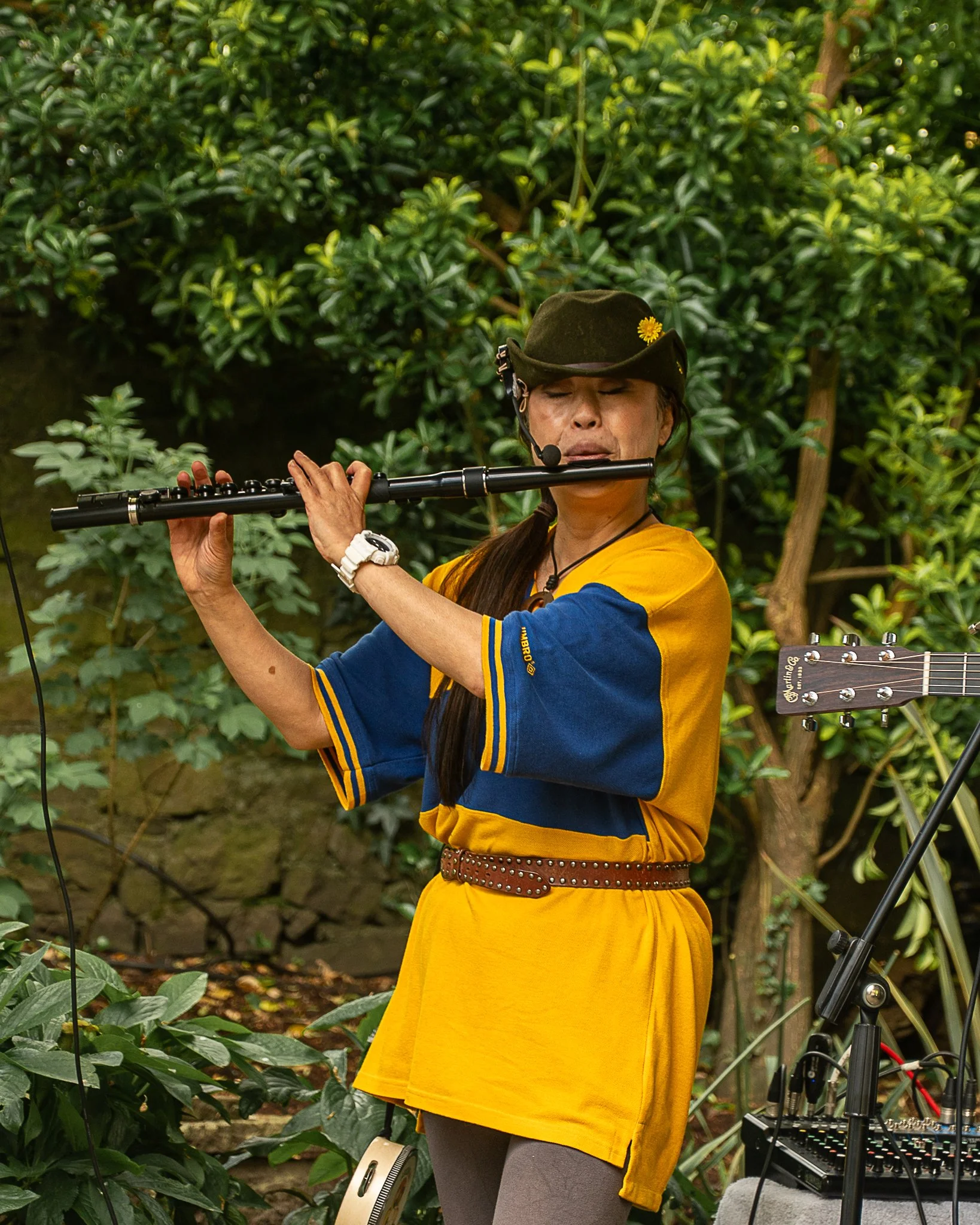 A woman wearing a yellow dress and gray leggings is playing a flute in a lush outdoor garden. She is wearing a dark green hat with a yellow flower and has a headset microphone. There is musical equipment, including a guitar and a keyboard, nearby.