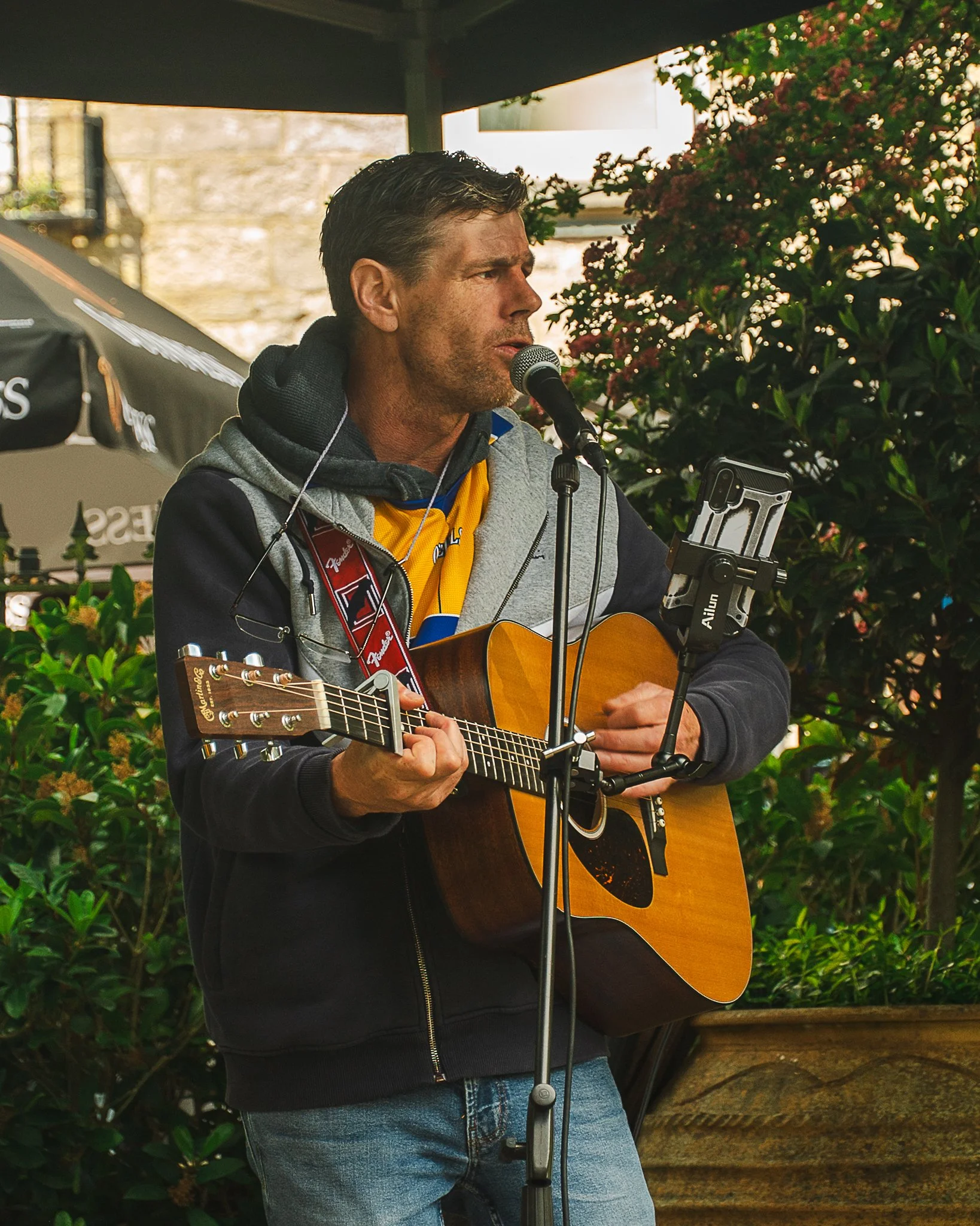 A man playing an acoustic guitar and singing into a microphone outdoors, surrounded by green foliage and pink flowers.
