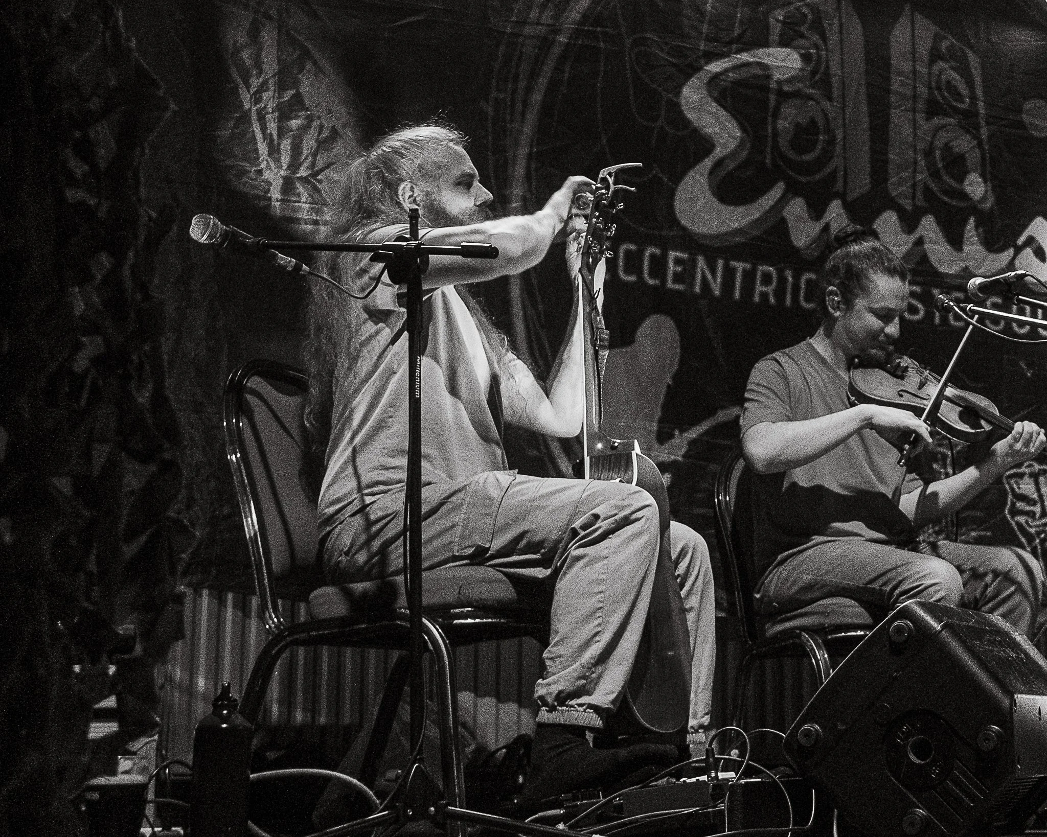 A person with long hair playing a violin on stage, with a microphone and another musician visible. The background features a large decorative banner or mural.
