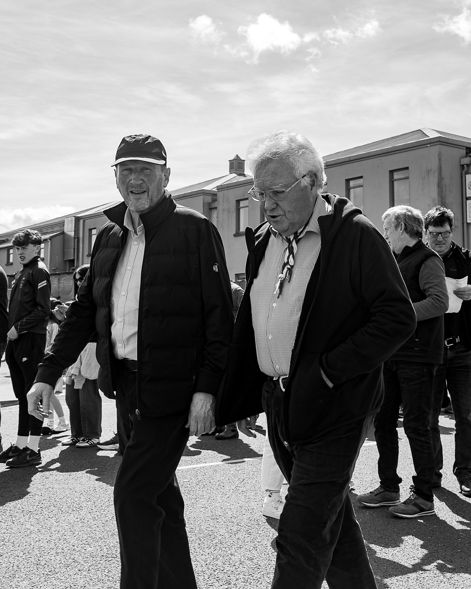 A group of people walking outdoors on a sunny day. The image features two older men in the foreground, one wearing a baseball cap and the other with glasses and white hair, both walking and engaged in conversation. There are other individuals in the 