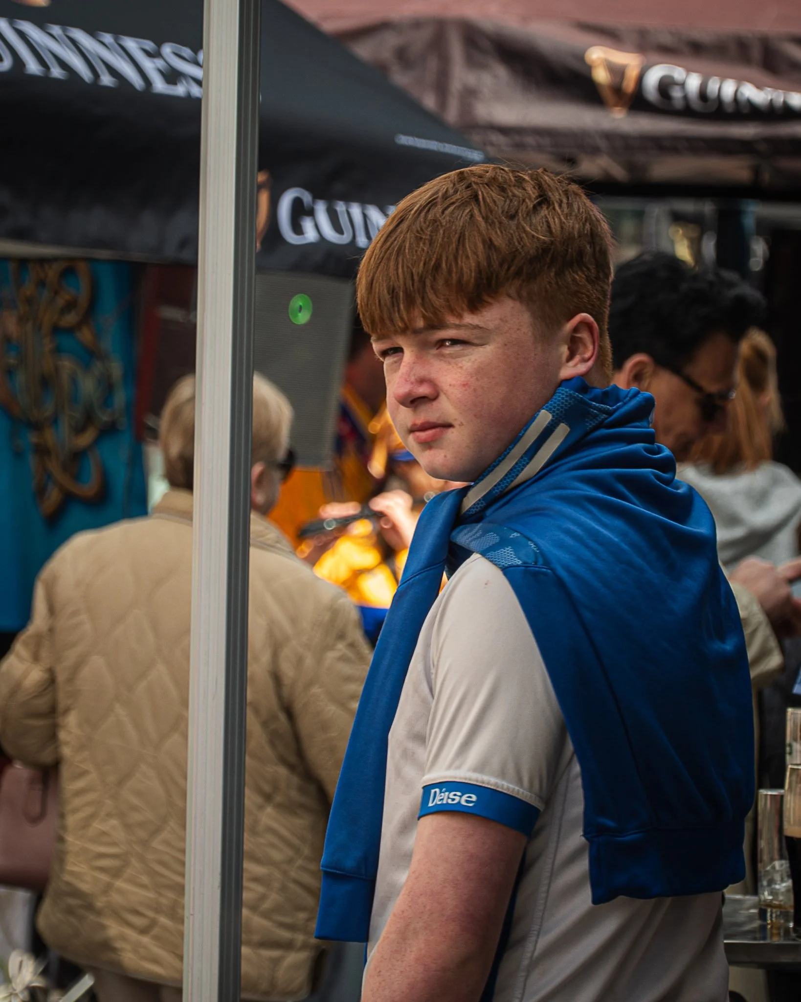 A young man with reddish hair wearing a blue and white sports shirt, standing in a busy indoor environment with many people around. He is seen from the side, with a focused expression.