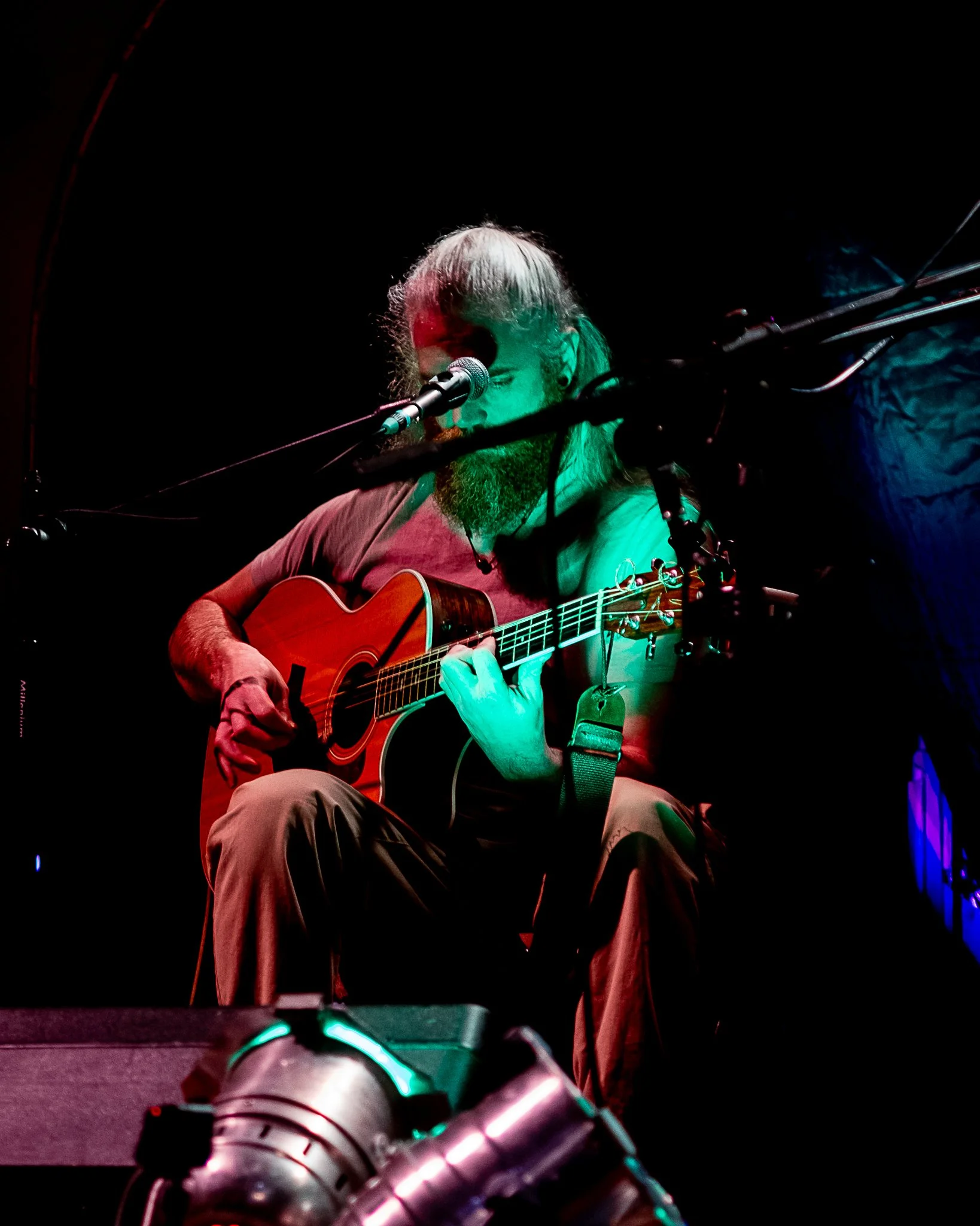 A man with long hair and a beard playing an acoustic guitar and singing into a microphone on a dark stage with colorful lighting.