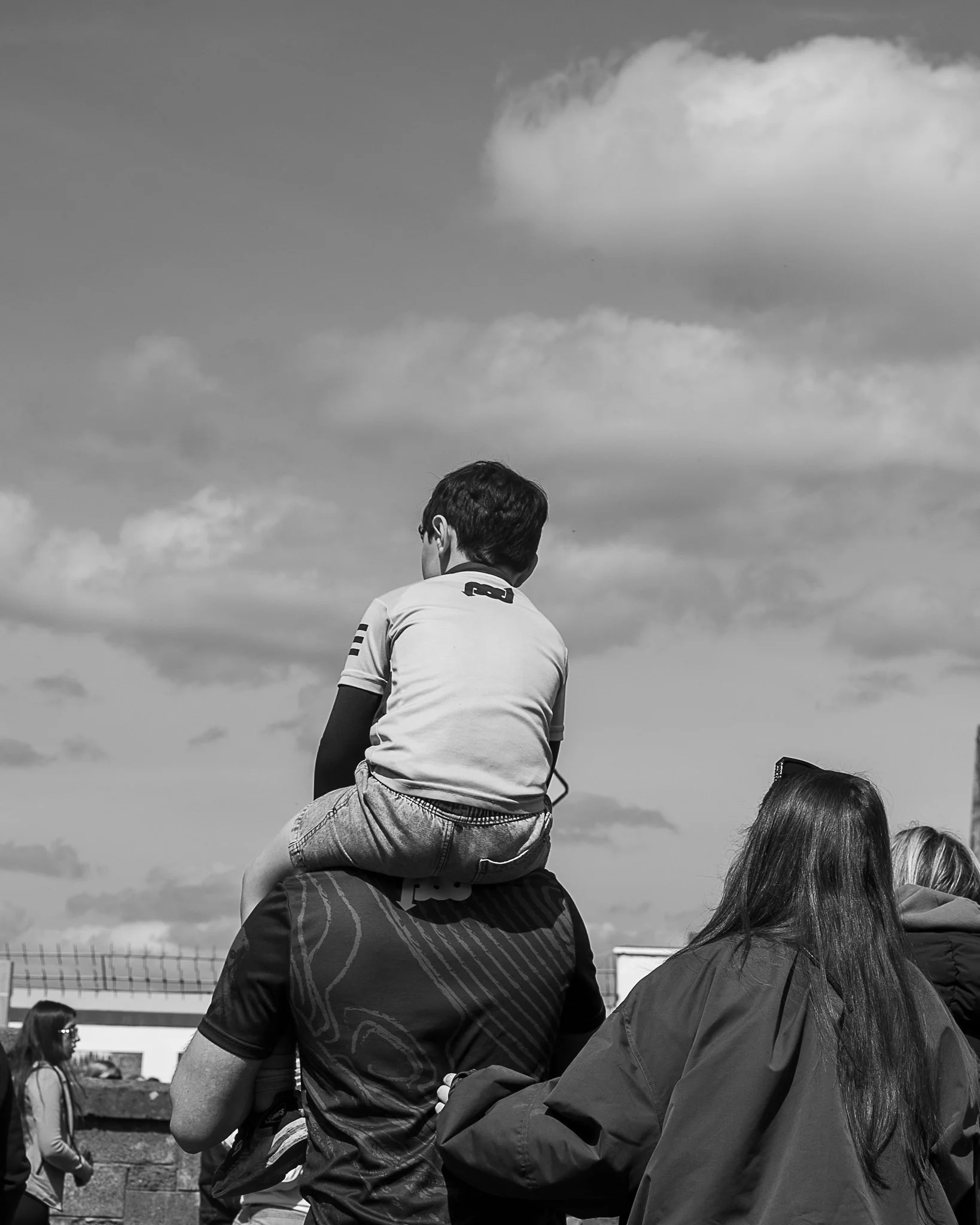 People outdoors in a sunny day, one person sitting on another person's shoulders, with a man and woman with long hair in the foreground, and a group of people in the background.