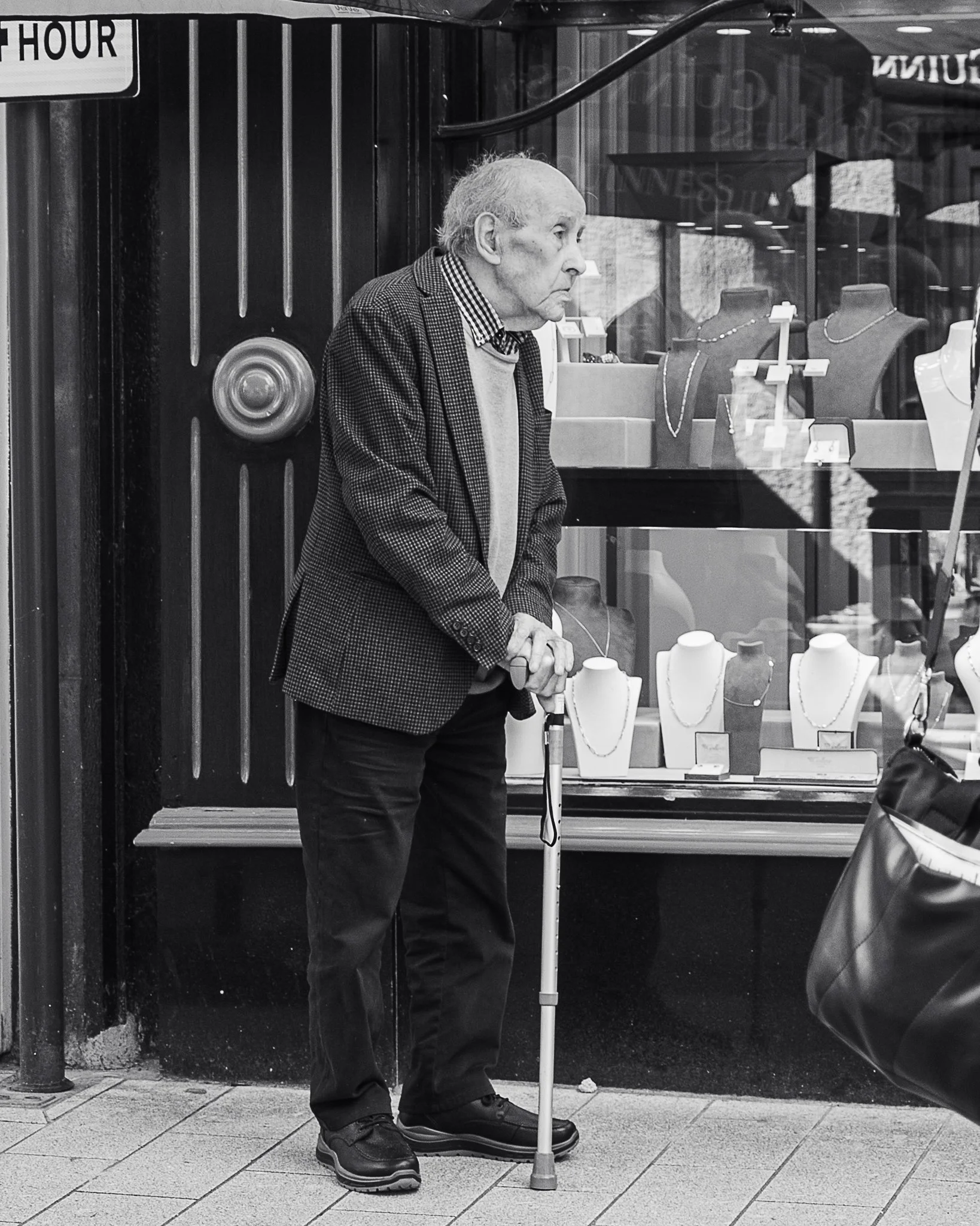 A black-and-white photo of an elderly man with thinning hair, standing on a sidewalk outside a jewelry store. He is wearing dark pants, a checkered blazer, a checkered shirt, and a sweater. He is using a white cane to assist his walking, and his gaze