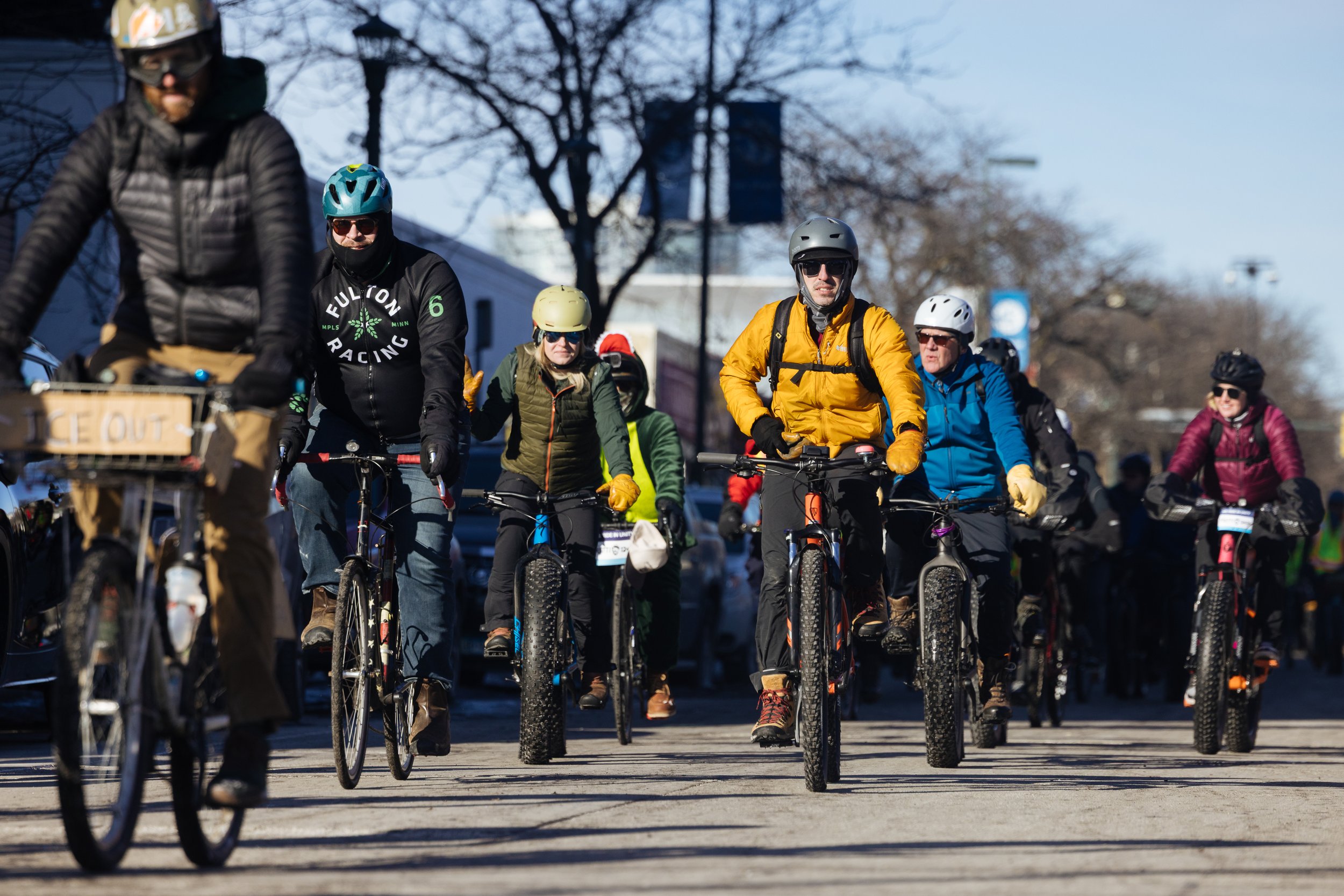 Group of people riding bicycles outdoors on a sunny day, some wearing helmets and sunglasses, with trees and buildings in the background.