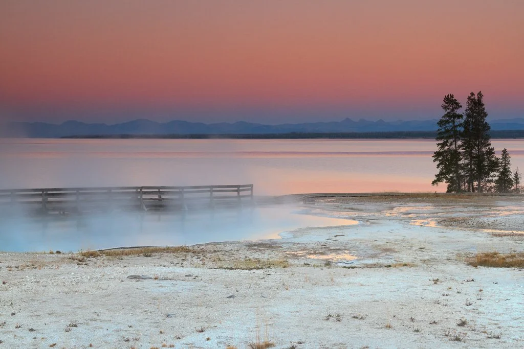 Steam rising from a geothermal hot spring, with a wooden dock extending into the water, a small group of trees, and a mountain range in the background during sunset.