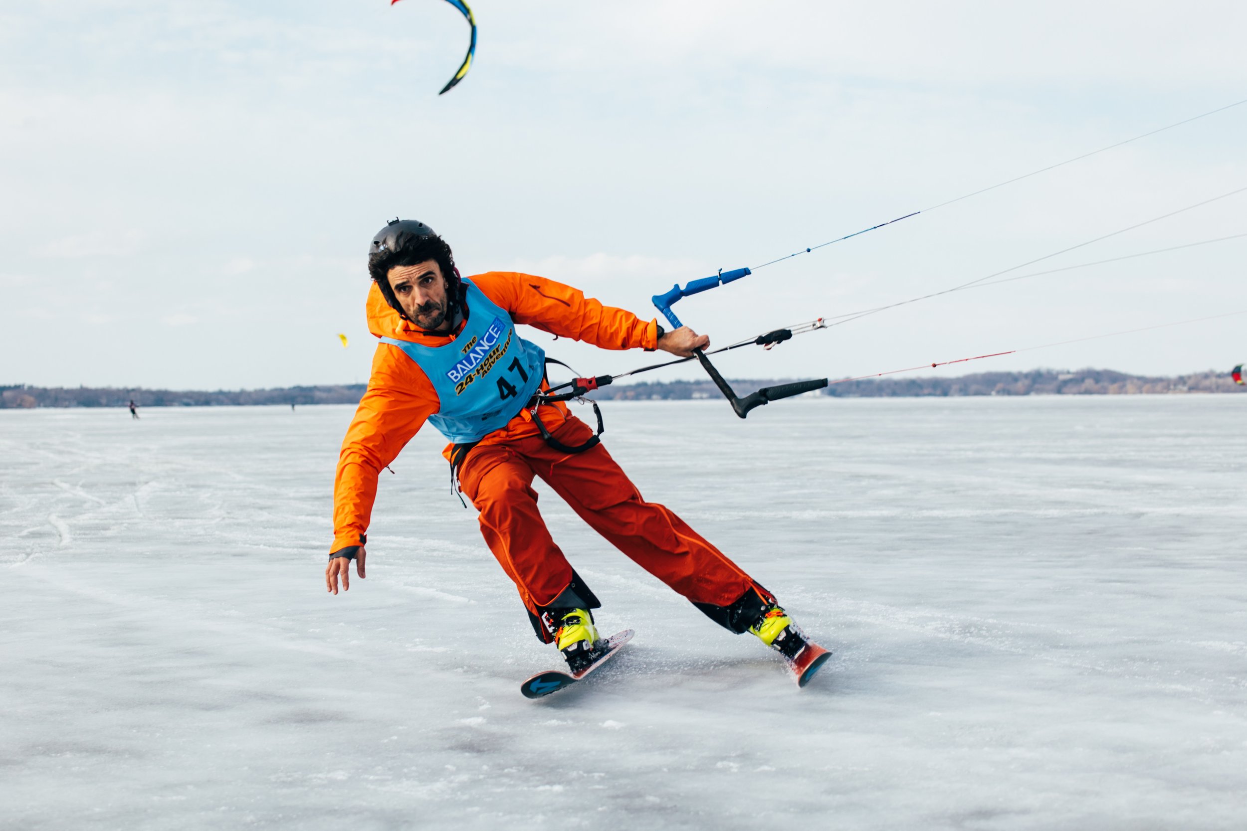 A man wearing a helmet, orange jacket, and orange pants snowboarding on a frozen lake with other kiteboarders in the background.