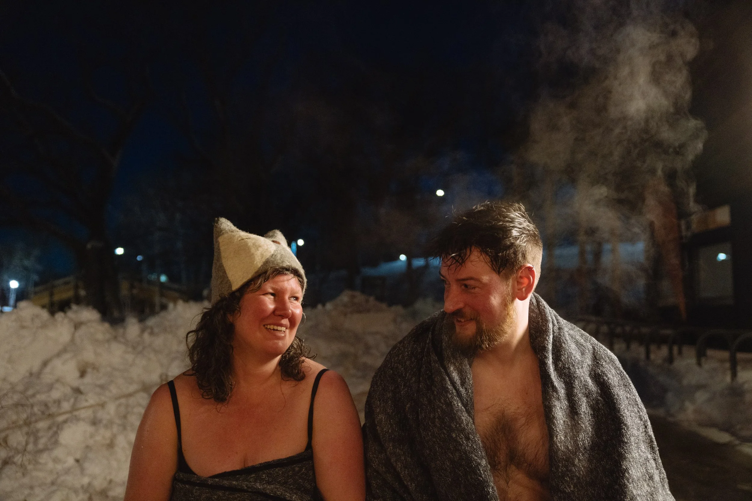 Two people, a woman and a man, are sitting outside at night, smiling and looking at each other. The woman is wearing a beige knit hat and a black spaghetti strap top. The man has a beard and is wearing a gray cardigan, and they appear to be enjoying 