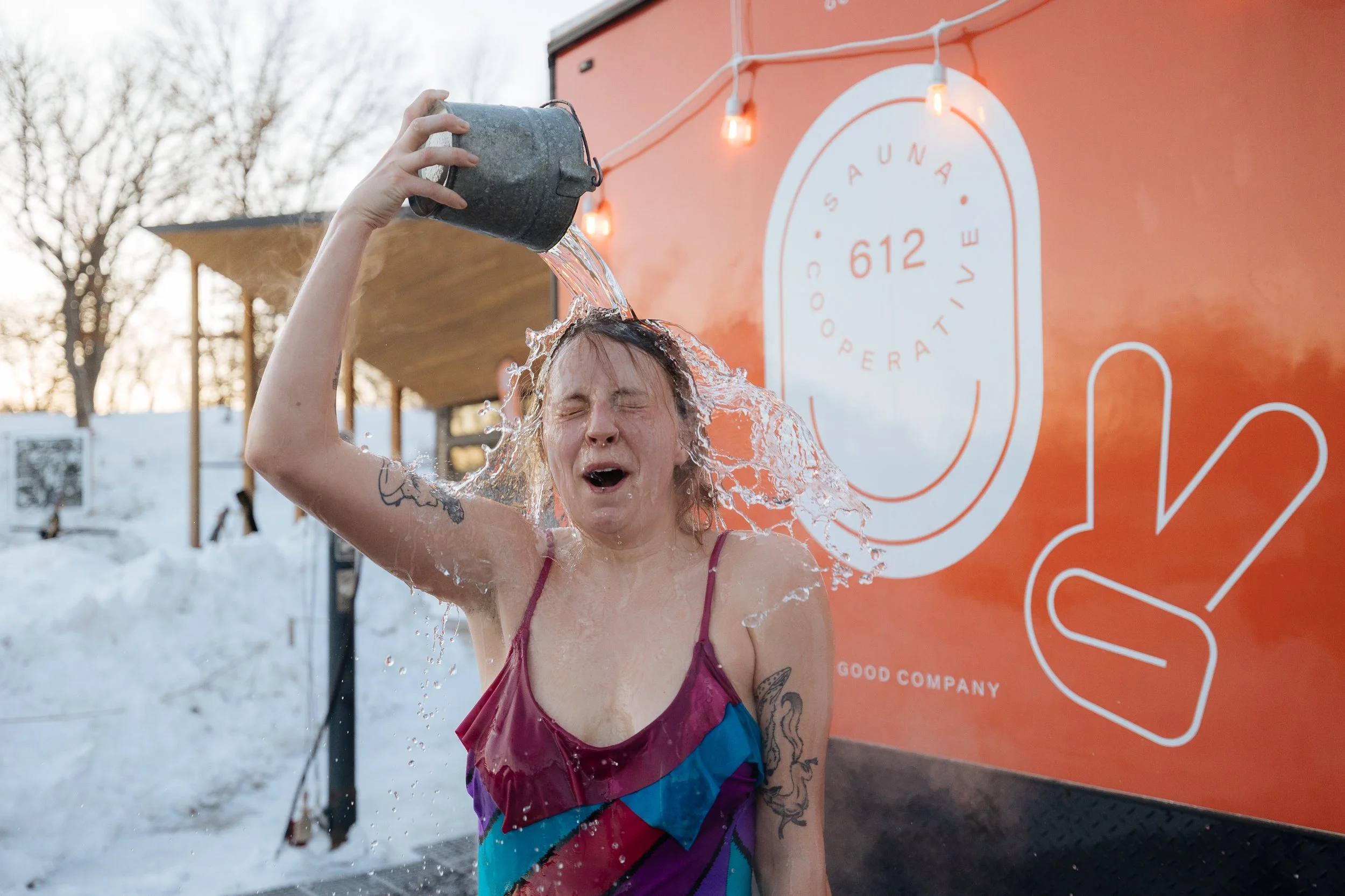 A person with tattoos on their arms and wearing a colorful dress is pouring water over their head at an outdoor event with snow on the ground. There is a large sign with the words 'OUSA' and a circular logo behind them, and string lights overhead.