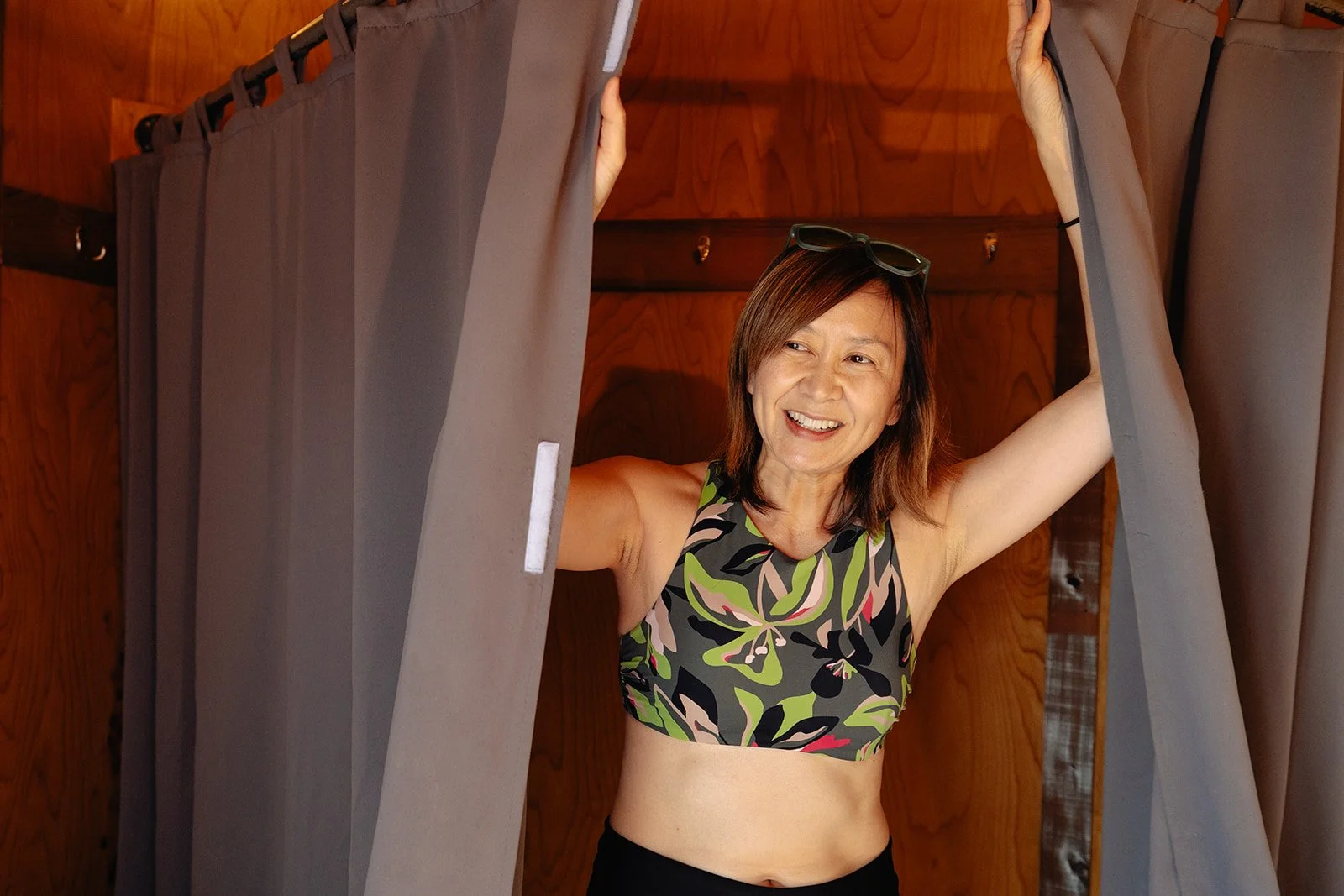 A woman smiling and laughing while standing behind a gray curtain in a wooden room, wearing a patterned sports bra.