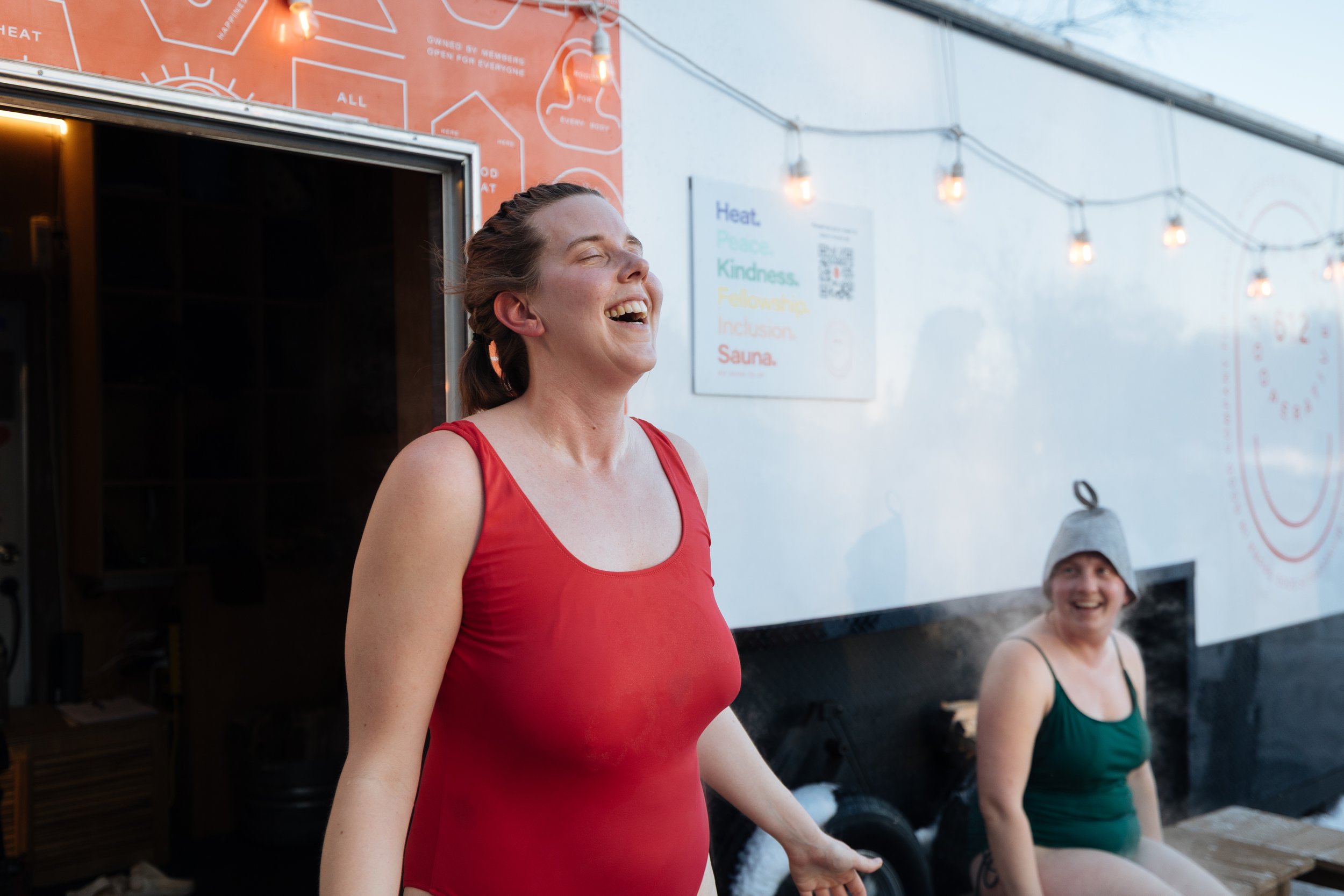A woman in a red tank top laughing outdoors at an event, with another woman in a green tank top and a gray beanie sitting in the background near string lights and signs.