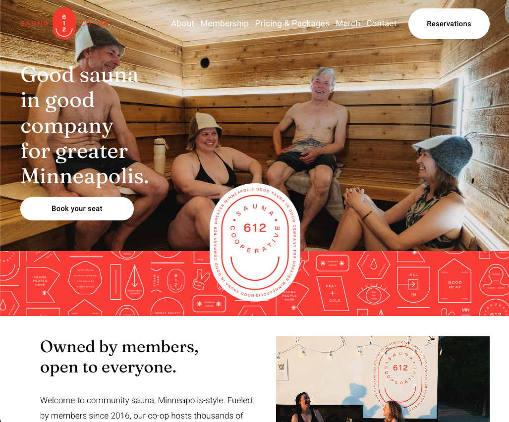 Group of four people inside a wooden sauna, smiling and relaxing on benches, wearing towels and sauna hats.