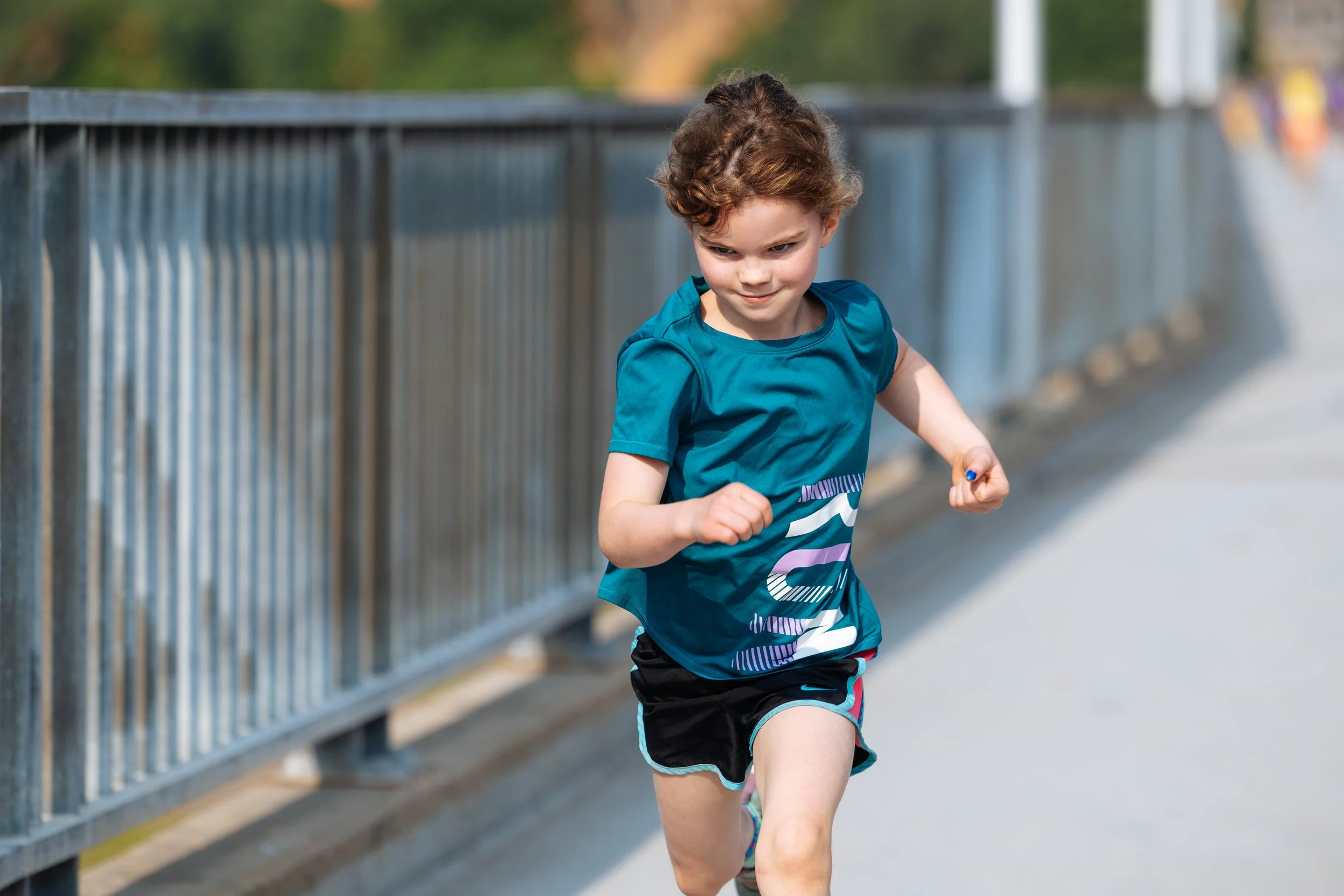 Young boy running outdoors on a pathway next to a metal railing, wearing a blue athletic shirt and shorts, with a concentration expression on his face.