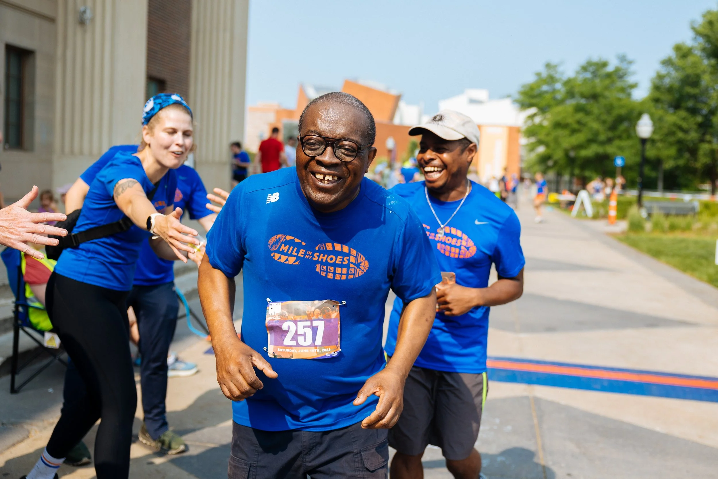 Smiling man with glasses and race bib running in a marathon, with other runners and spectators behind him on a city street on a sunny day.